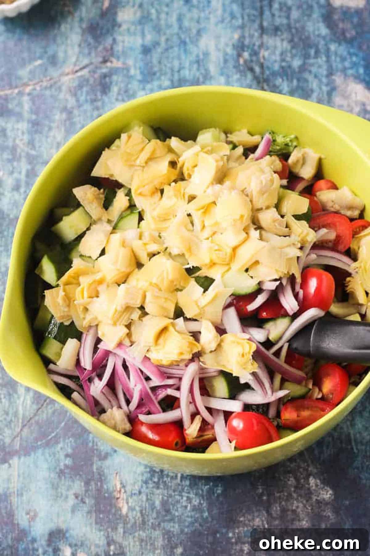 Zesty Lemon & Artichoke Pasta Salad Recipe for Summer 4 Chopped artichokes, tomatoes, cucumbers, and sliced red onion in a green mixing bowl.