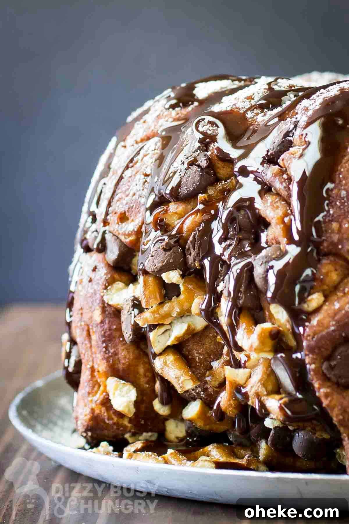Chocolate Pretzel Biscuit Monkey Bread Recipe 2 Side shot of whole monkey bread sprinkled with powdered sugar and chocolate syrup on a white plate on a wooden table.