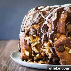 Chocolate Pretzel Biscuit Monkey Bread Recipe 12 Side shot of whole monkey bread sprinkled with powdered sugar and chocolate syrup on a white plate on a wooden table.