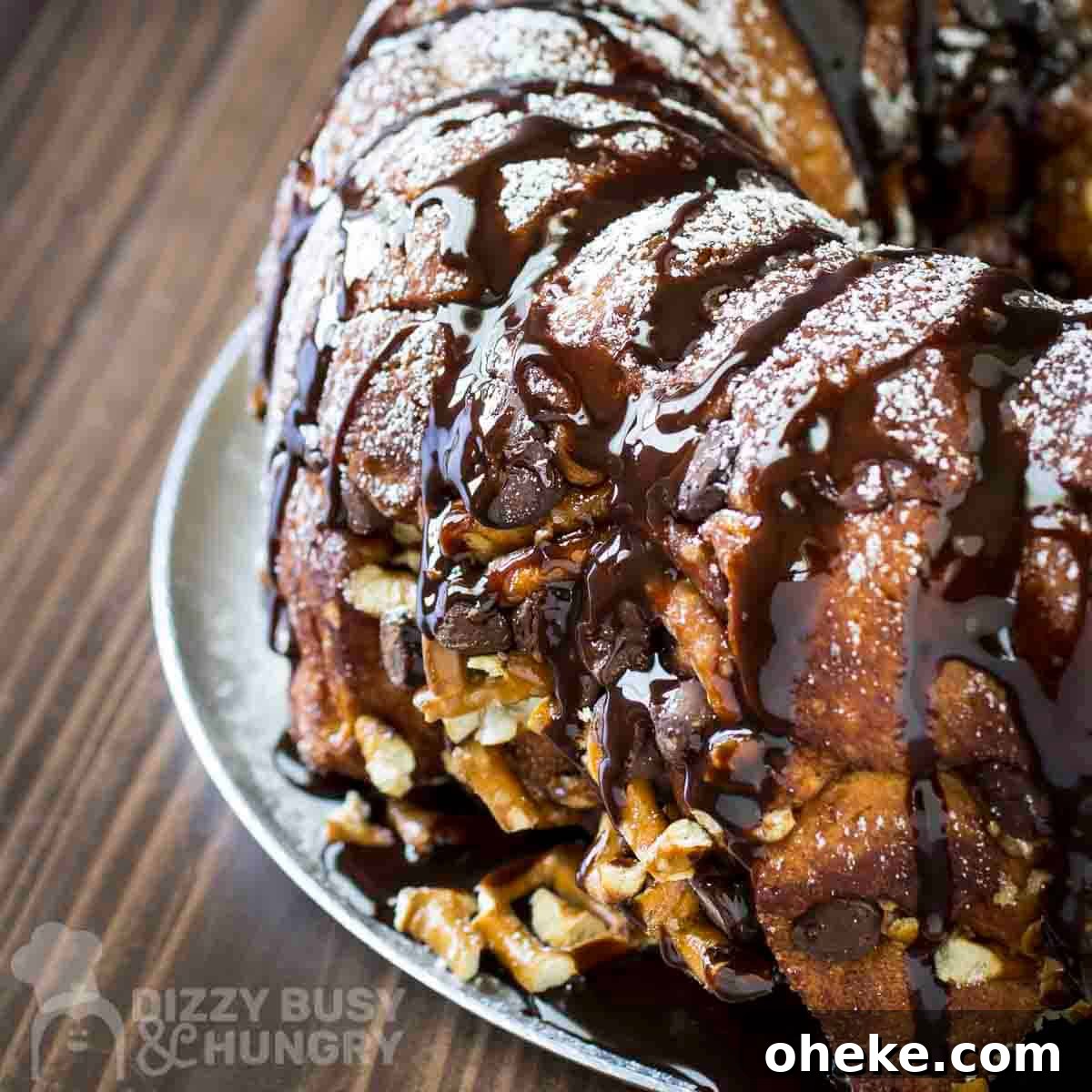 Chocolate Pretzel Biscuit Monkey Bread Recipe 10 Side shot of whole monkey bread sprinkled with powdered sugar and chocolate syrup on a white plate on a wooden table.