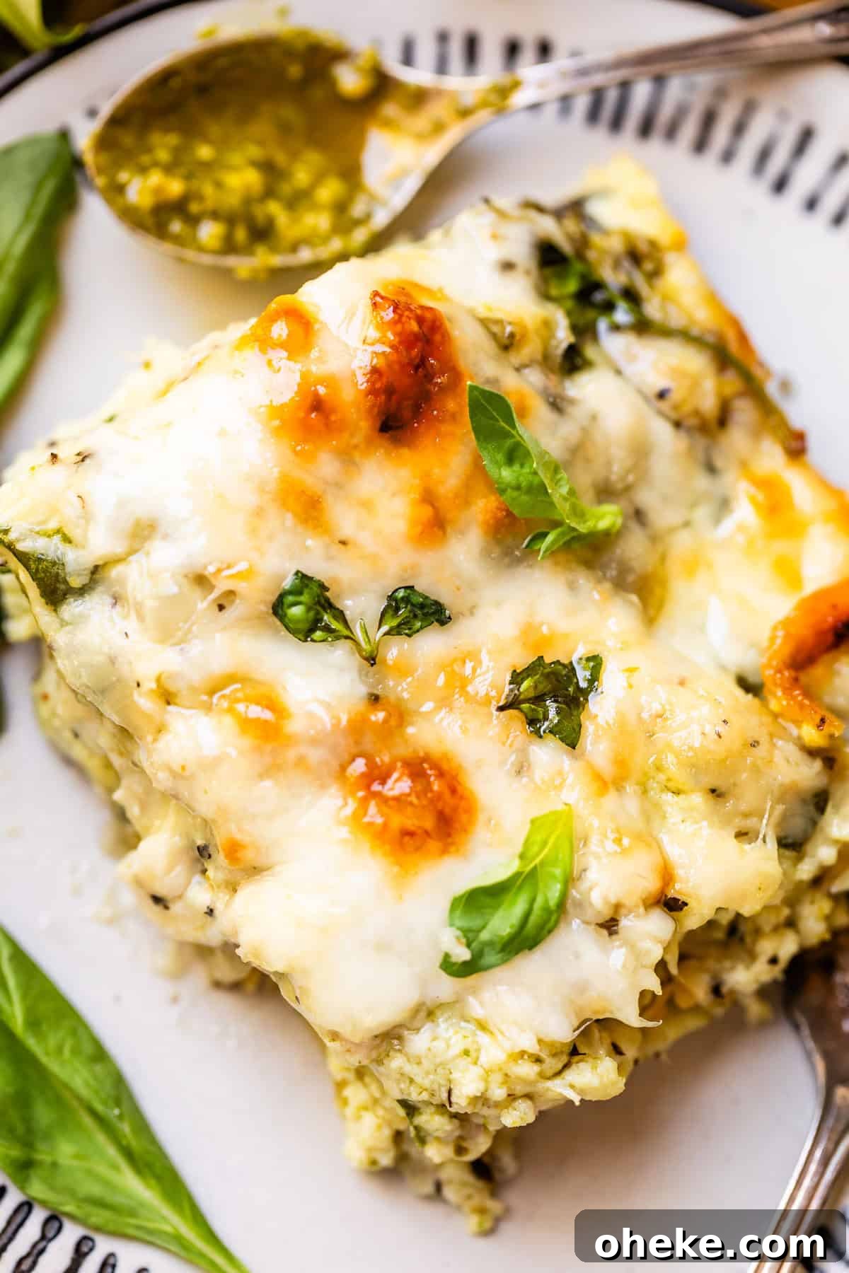 A close-up of a slice of cheesy lasagna topped with melted cheese and fresh basil leaves on a white plate, with a spoonful of green pesto sauce in the background.