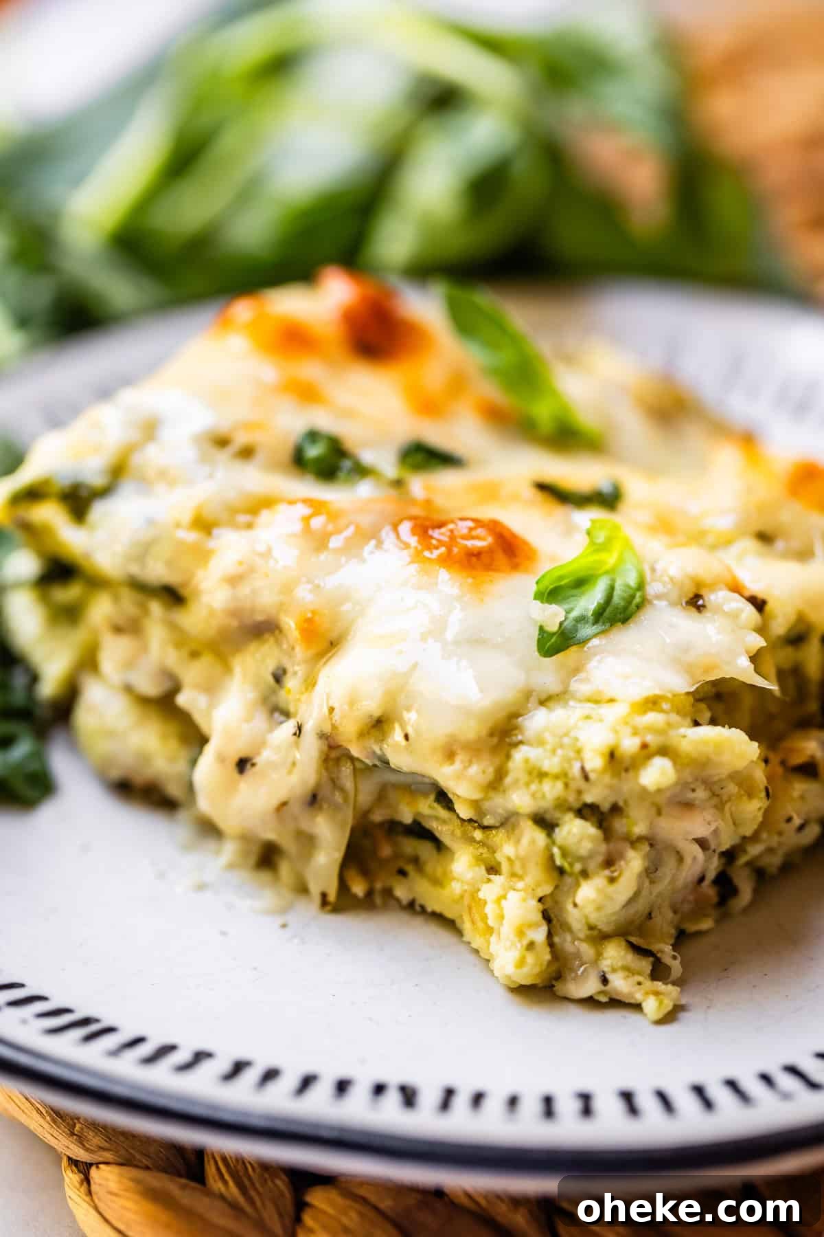 A close-up of a slice of creamy, cheesy vegetable lasagna garnished with fresh basil, served on a white plate with a decorative rim. Green leafy vegetables are blurred in the background.