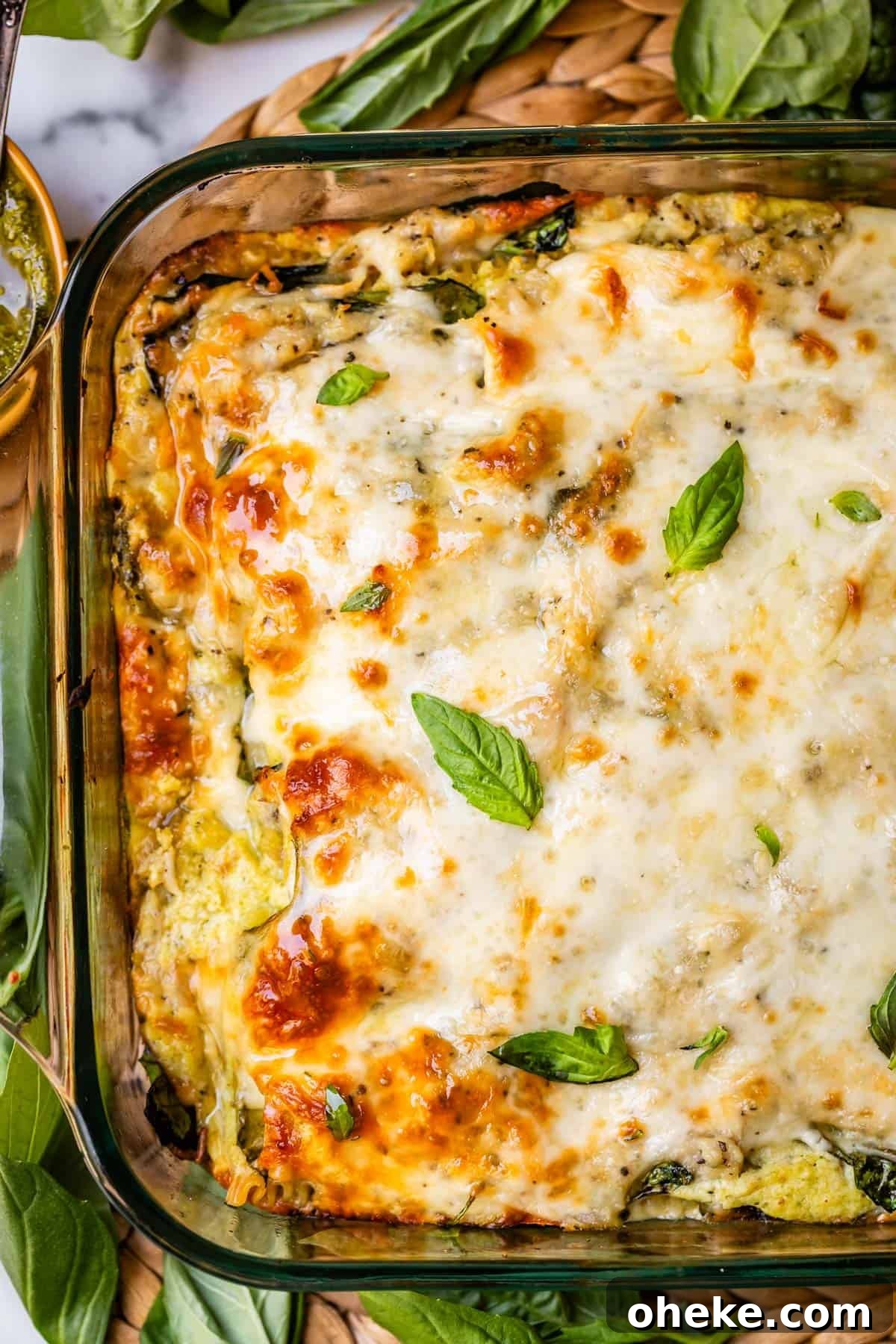 A close-up of a baked cheesy casserole in a glass dish, topped with melted golden cheese and fresh basil leaves, surrounded by fresh spinach leaves.