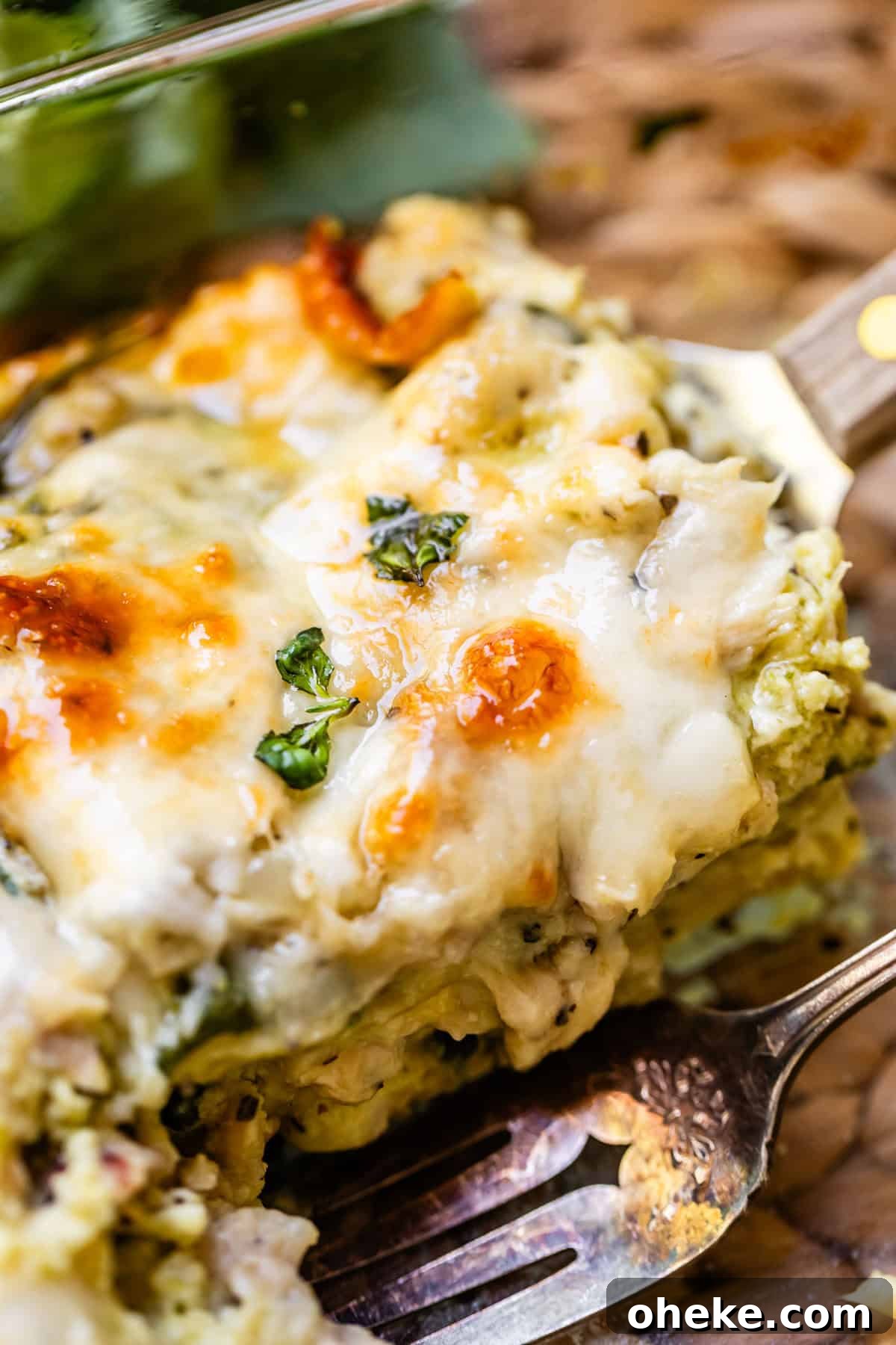 A close-up of a cheesy, golden-brown slice of vegetable lasagna on a fork, topped with melted cheese and herbs, showing creamy layers and a slightly crisp surface.