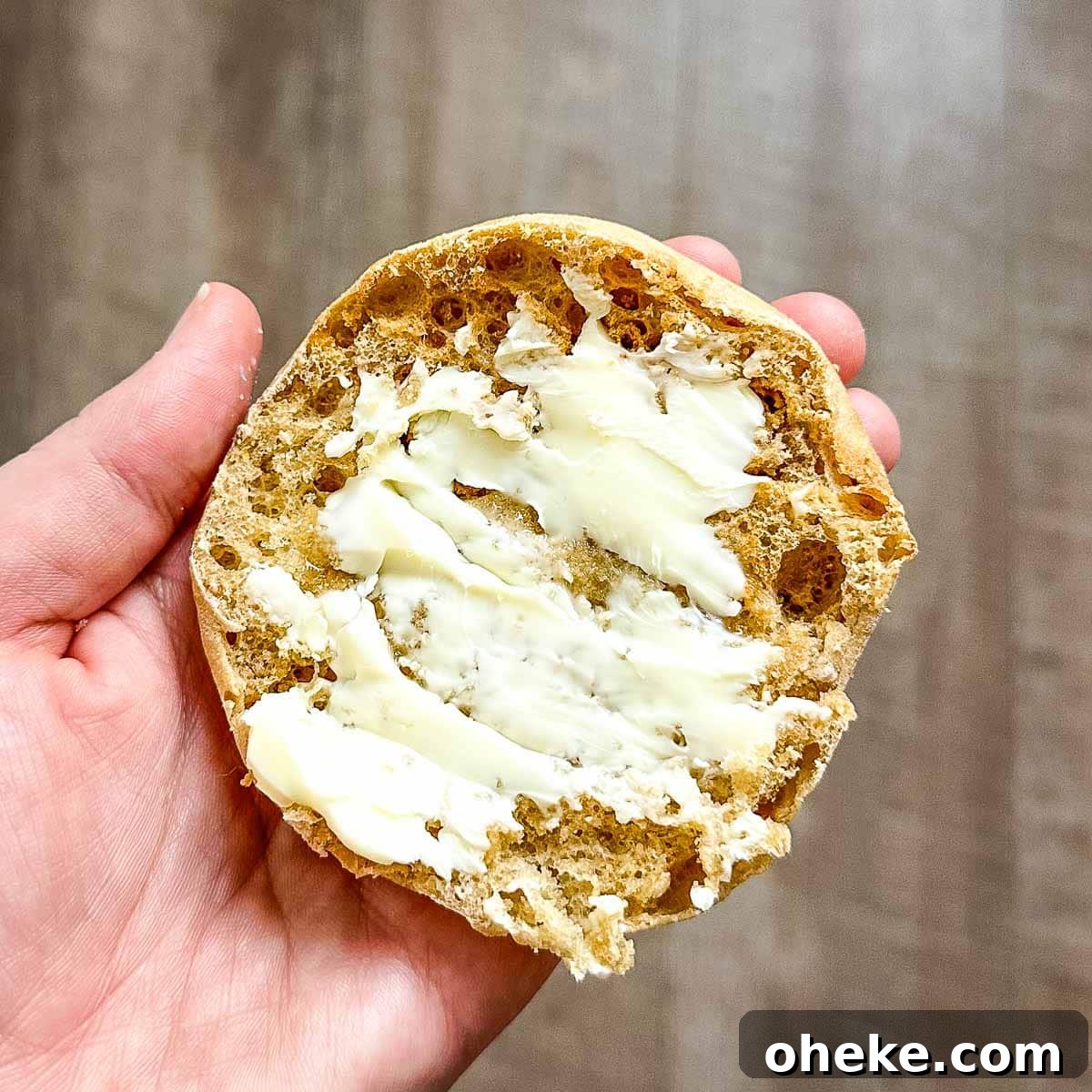 A buttered English muffin is held over a wooden backdrop.