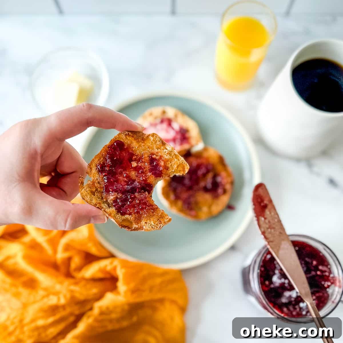 An English muffin with a bite taken out of it is held over a scene with a blue plate, a yellow linen, jam, a knife, coffee, and orange juice.