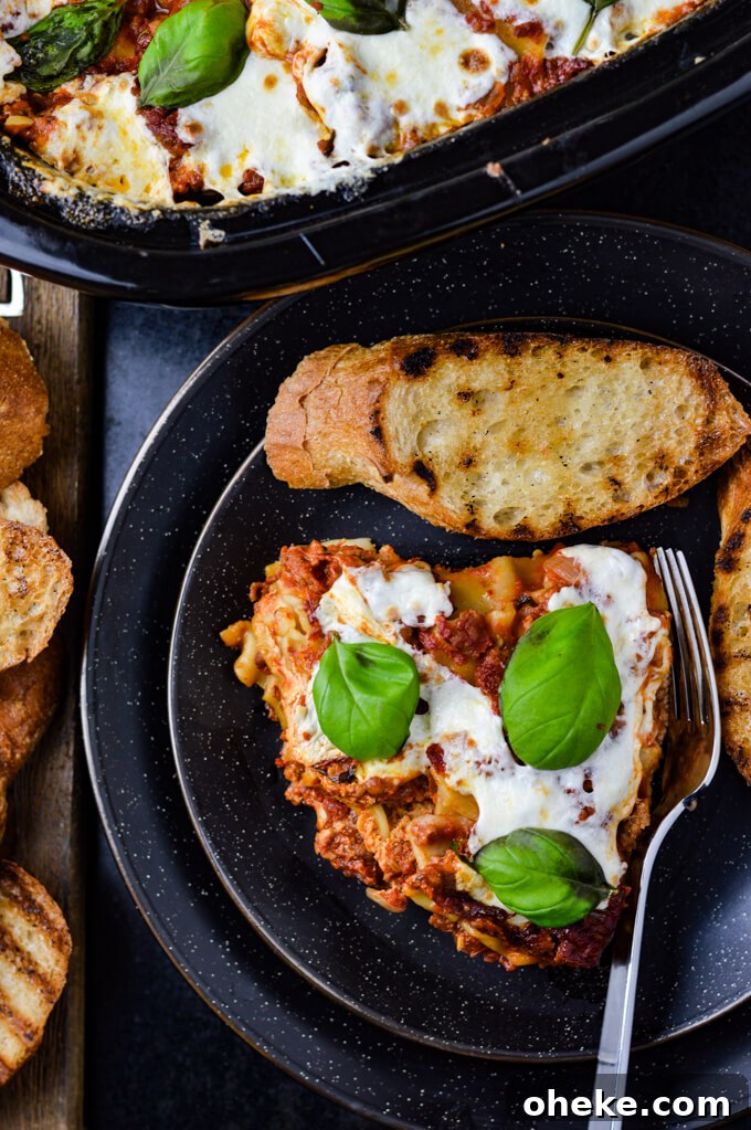 Slow-Cooker Lasagna Recipe for Tender, Cheesy Layers 13 A slice of homemade lasagna with fresh mozzarella and basil leaves on top sits on a black pate with toasted slices of toasted garlic bread. The pan of lasagna sits in the corner of photo.