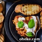 A slice of homemade lasagna with fresh mozzarella and basil leaves on top sits on a black pate with toasted slices of toasted garlic bread. The pan of lasagna sits in the corner of photo.