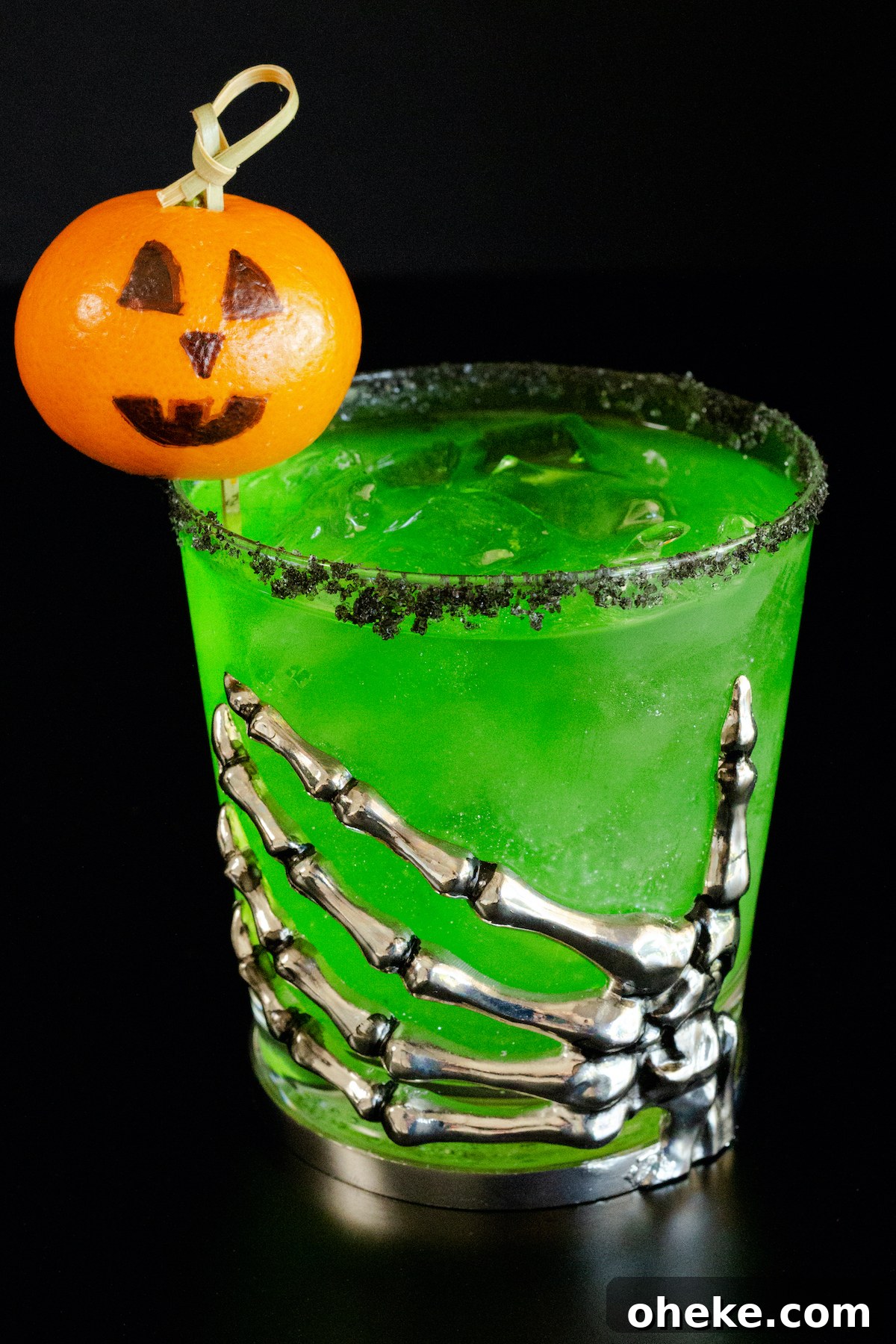 A lowball glass with a skeleton hand on the front is filled with a bright green margarita. There is a black salt rim and jack o lantern garnish.