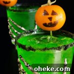 Two bright green Halloween margaritas with black salt rims and mandarin oranges with jack-o-lantern faces drawn on as garnishes sit on a black background.