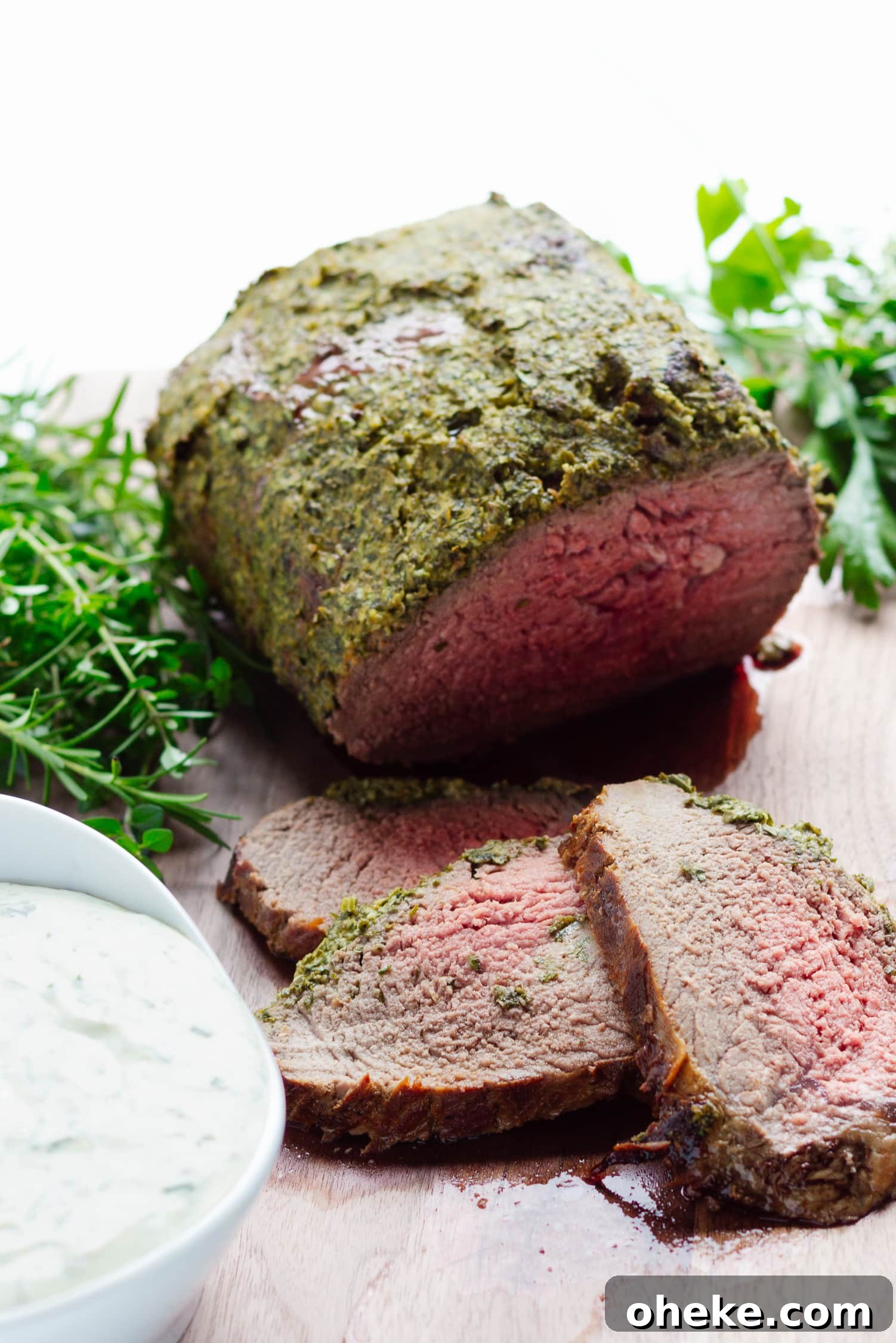 Herb Crusted Roast Beef on a wooden cutting board with 3 slices carved off in front.  Also a bowl of creamy herb sauce and fresh herbs on the board.