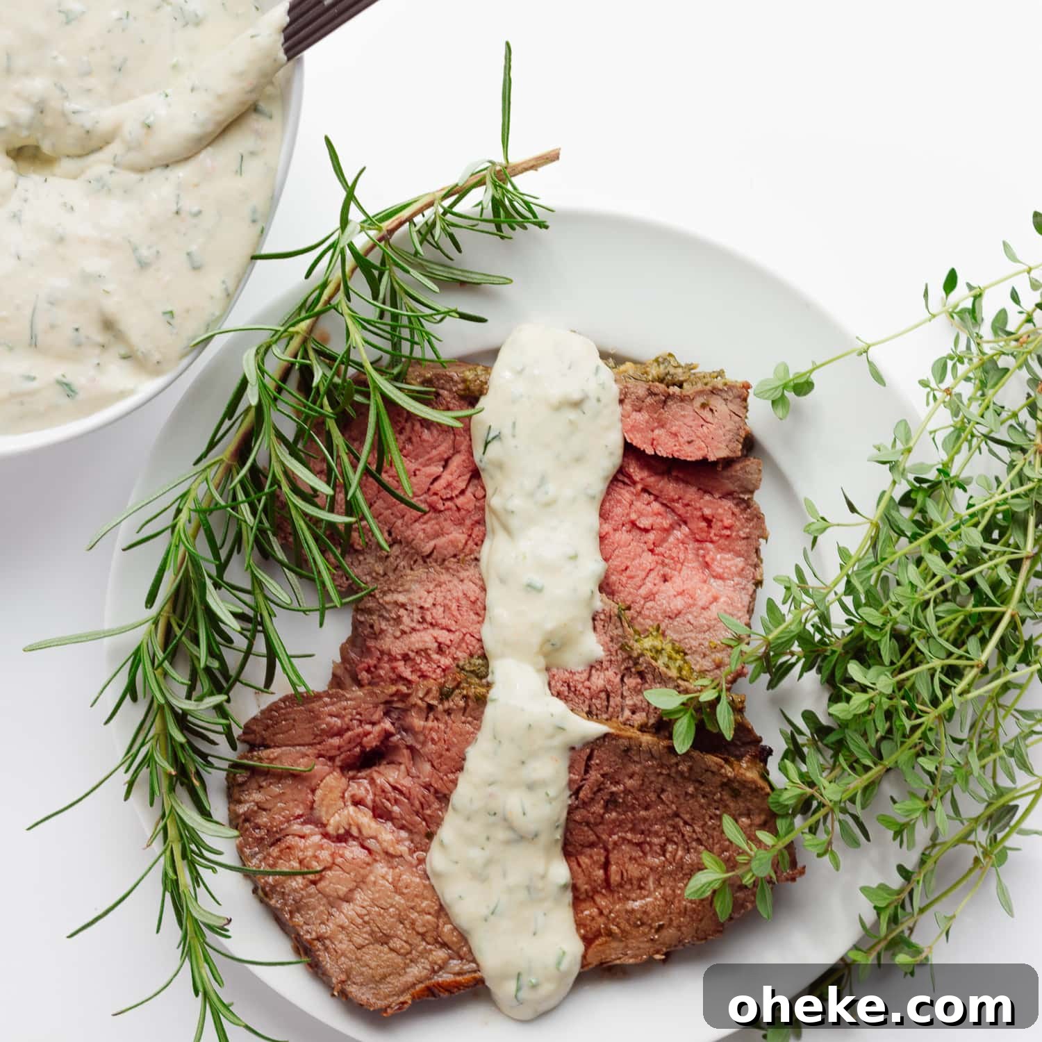 Slices of roast beef on a white plate with a drizzle of creamy herb sauce down the middle.  There is also a bowl of the same sauce in the corner and a sprig of rosemary and thyme on each side of the plate.