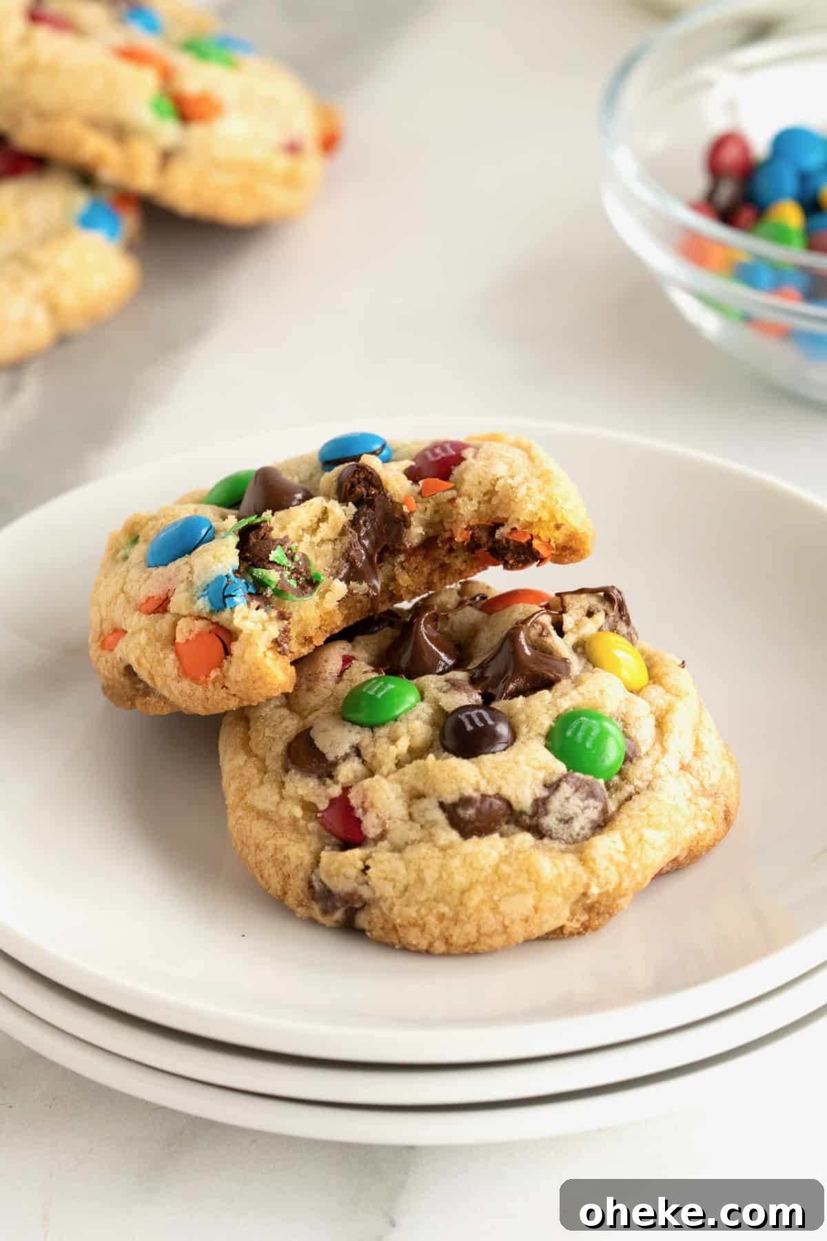 Two chocolate chip M&M cookies on a white plate and one has a bite out of it.