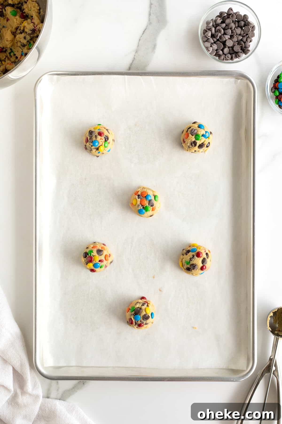 Unbaked balls of M&M chocolate chip cookie dough on a parchment lined baking sheet.