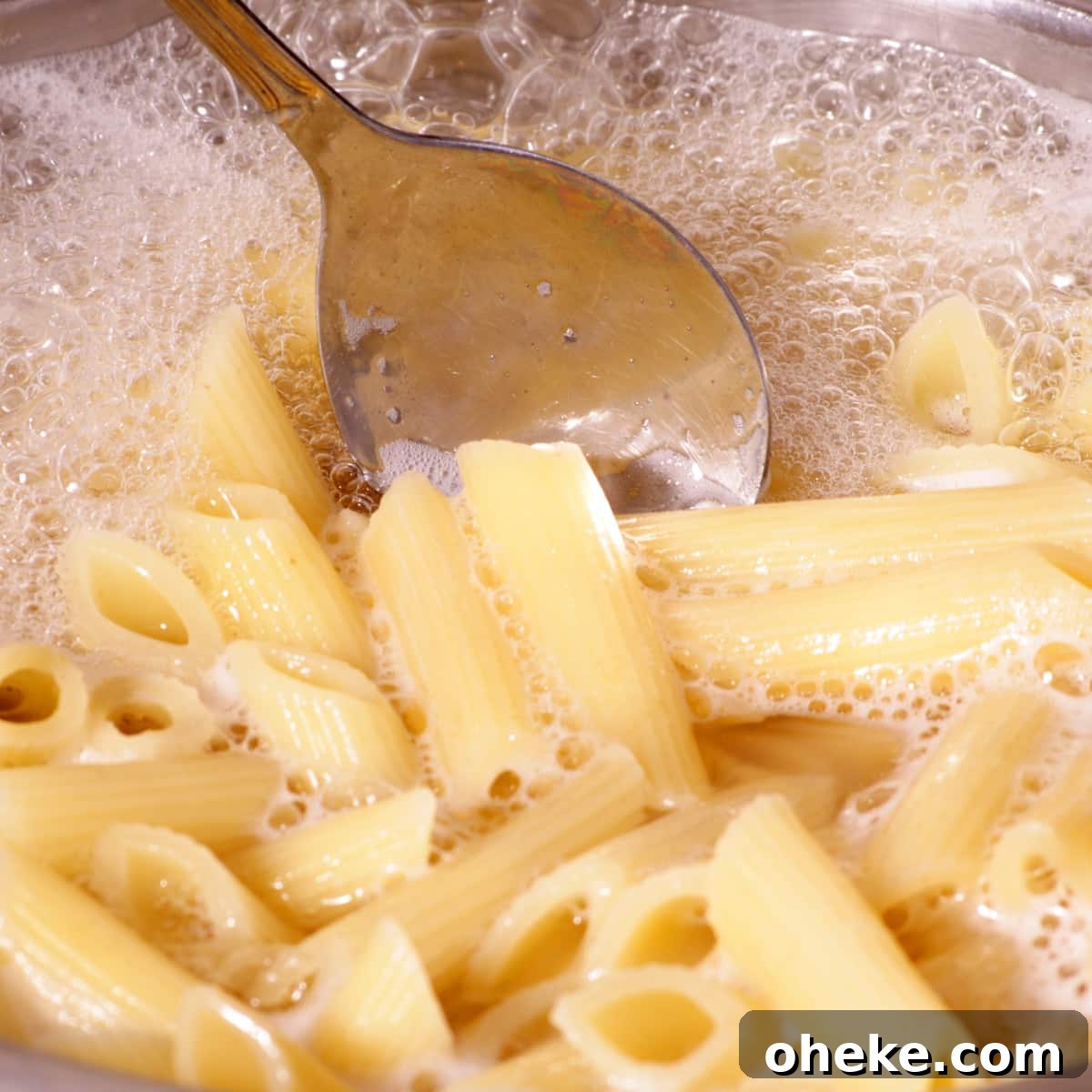 Perfect Pasta: Mastering Water, Salt and Timing 2 Penne pasta in a stainless steel pot of boiling water with a silver spoon stirring the noodles.