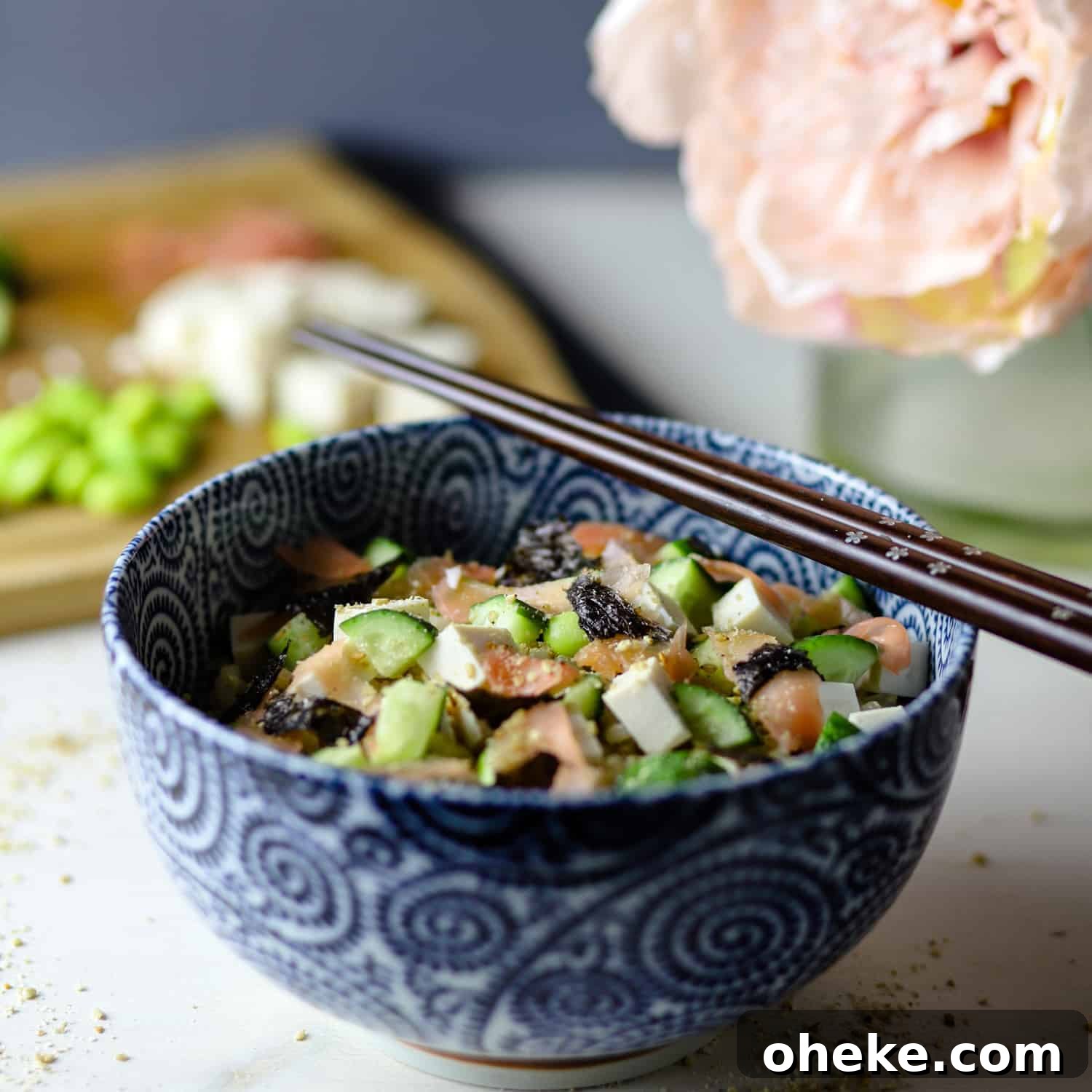Photo of white marble counter with blue decorated bowl filled with sushi rice and vegan toppings (tofu, cucumber, seaweed, sushi ginger, etc) and flowers and ingredients on a chopping board in the background.