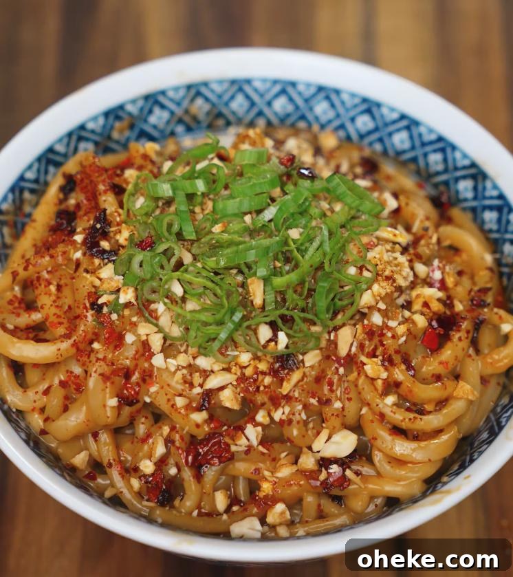 Spicy Peanut Noodles with Garlic and Chili Oil 2 A close-up shot of spicy peanut butter noodles topped with chopped scallions, crushed peanuts, chili flakes, and chili oil, served in a patterned blue and white bowl on a wooden table.