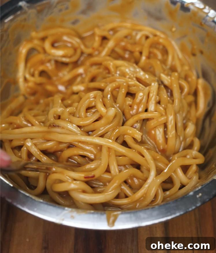 Spicy Peanut Noodles with Garlic and Chili Oil 5 Cooked udon noodles are being mixed with the spicy peanut butter sauce in a large bowl until thoroughly coated, creating a glossy and rich noodle base before garnishing.