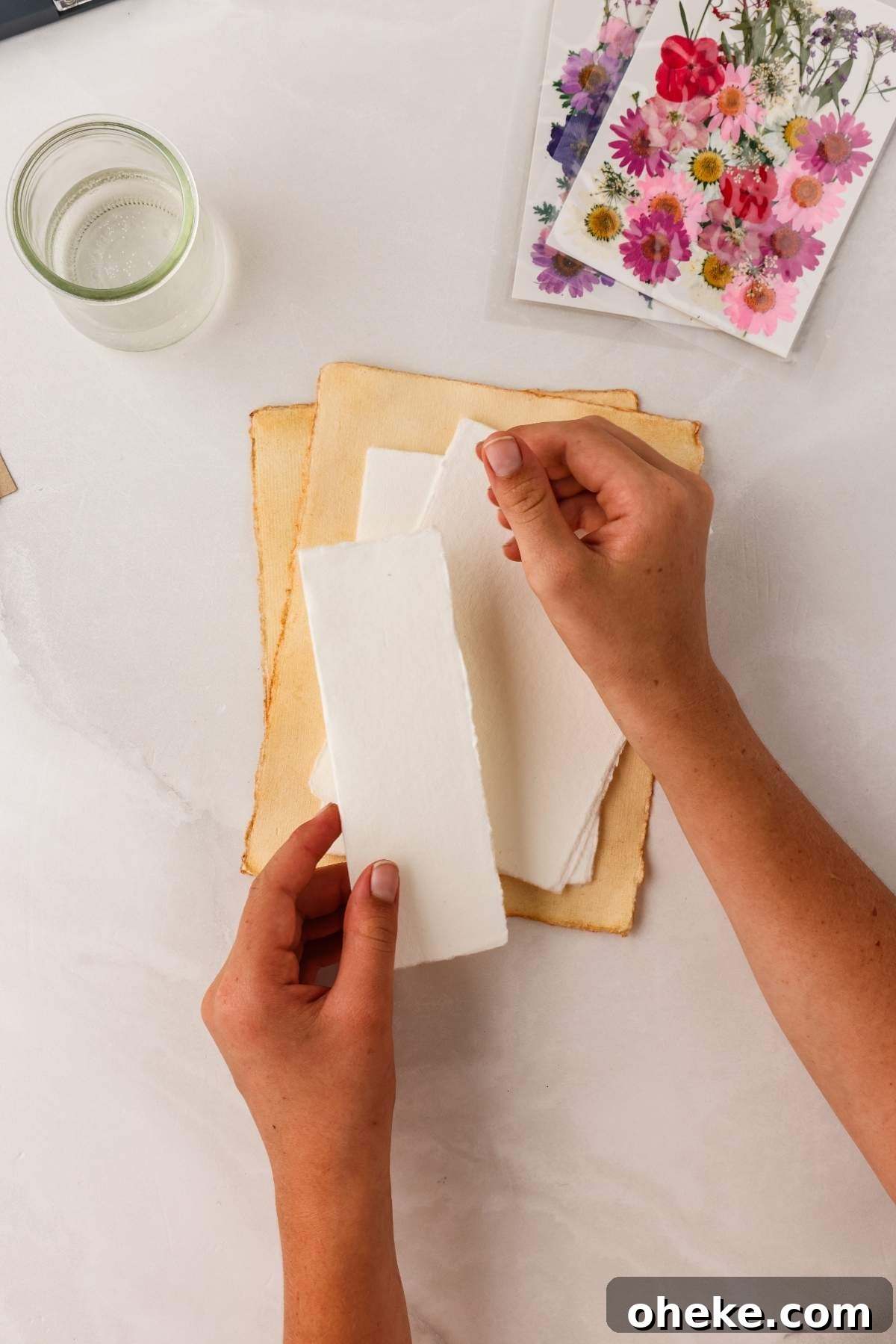 DIY Pressed Flower Bookmarks: Step-by-Step Craft Tutorial 12 Hands holding a piece of paper over a stack of papers with a water glass and floral cards nearby, perfect for crafting pressed flower bookmarks.