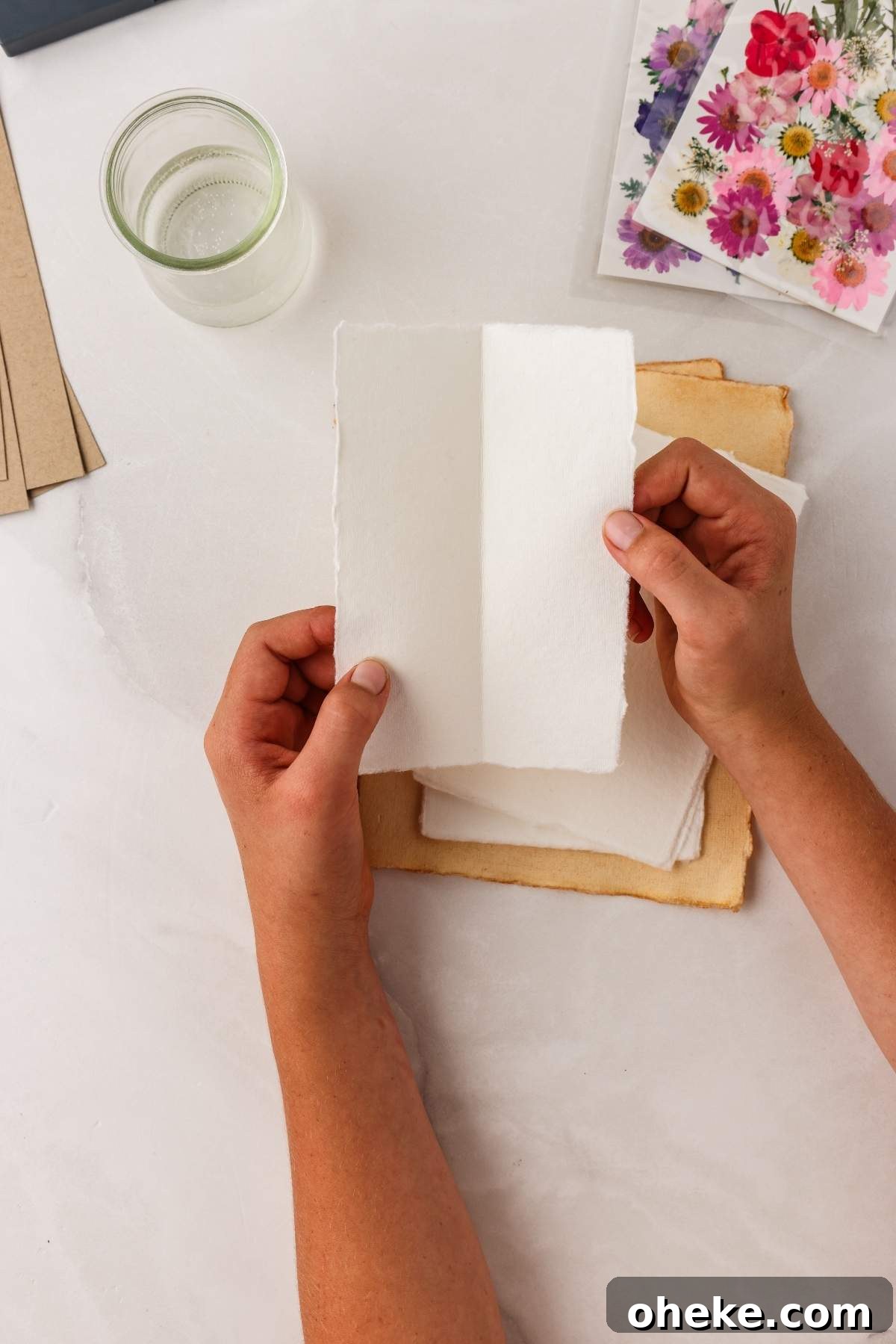 DIY Pressed Flower Bookmarks: Step-by-Step Craft Tutorial 13 Hands holding a blank piece of paper above a stack of papers, with a jar of water and floral stickers nearby on a white surface—perfect for crafting pressed flower bookmarks DIY projects.