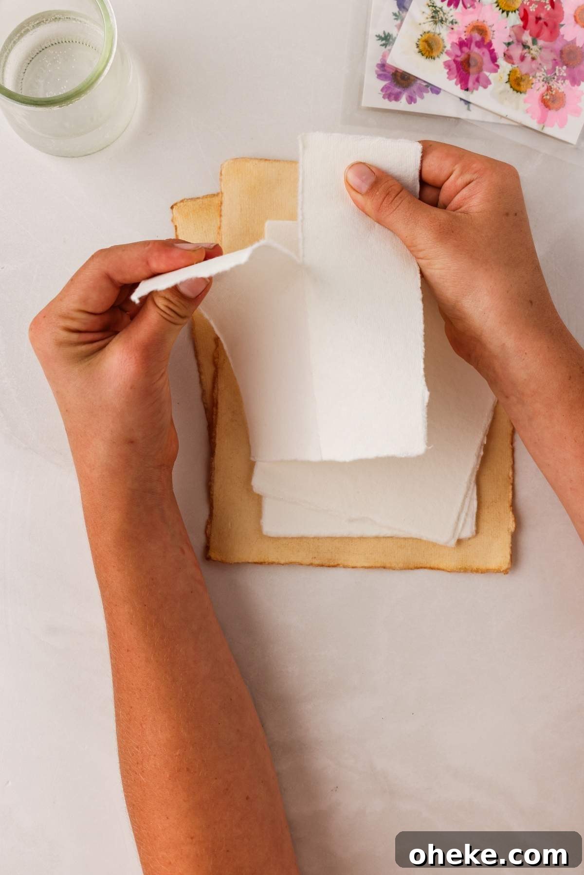 DIY Pressed Flower Bookmarks: Step-by-Step Craft Tutorial 14 Hands tearing a piece of white paper over a stack of aged papers, with pressed flower bookmarks nearby, and a glass of water alongside vibrant flower stickers.
