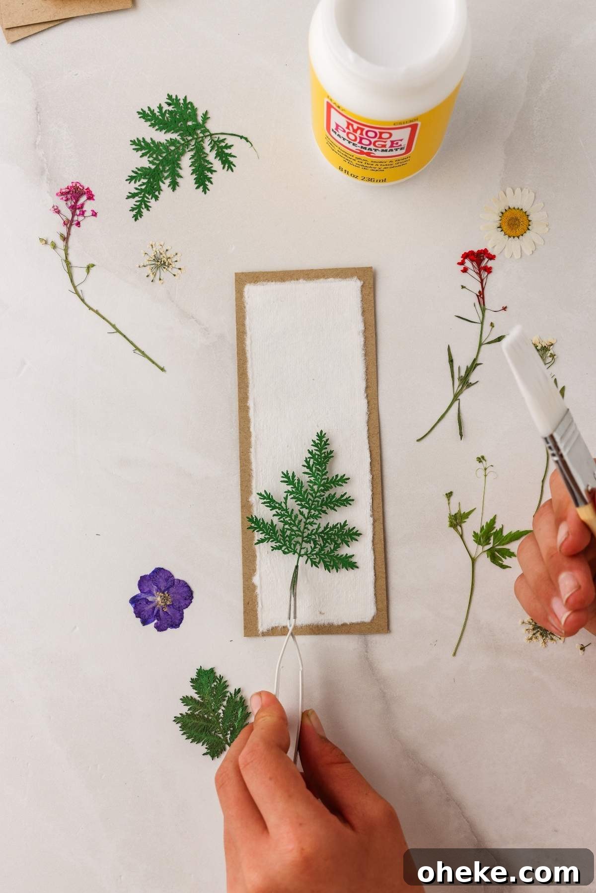 DIY Pressed Flower Bookmarks: Step-by-Step Craft Tutorial 15 Person crafting a DIY pressed flower bookmark, using glue and a paintbrush to carefully arrange dried flowers on a white rectangular card.