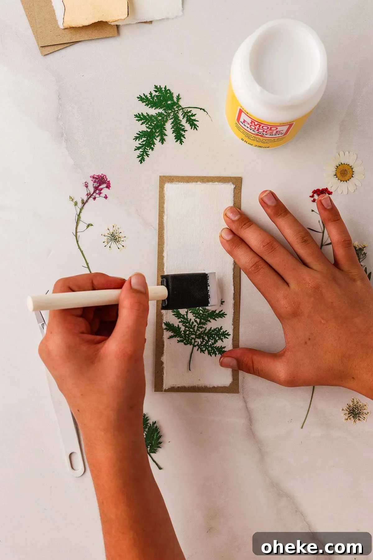 DIY Pressed Flower Bookmarks: Step-by-Step Craft Tutorial 16 A person creating a pressed flower bookmark DIY, gluing delicate blooms onto paper with a brush and Mod Podge on a marble surface.