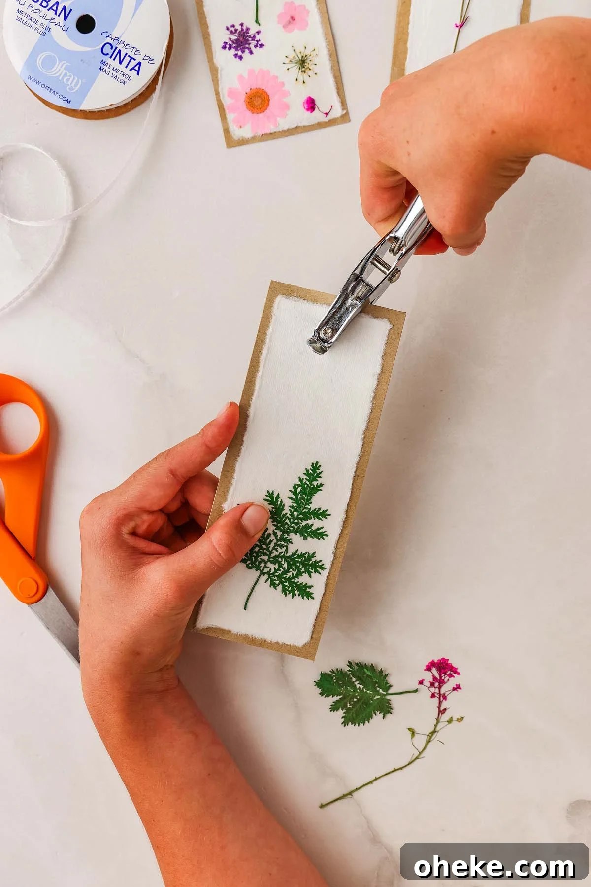 DIY Pressed Flower Bookmarks: Step-by-Step Craft Tutorial 17 Hand uses hole punch on a pressed flower bookmark DIY, decorated with delicate leaves; scissors and ribbon rest nearby.