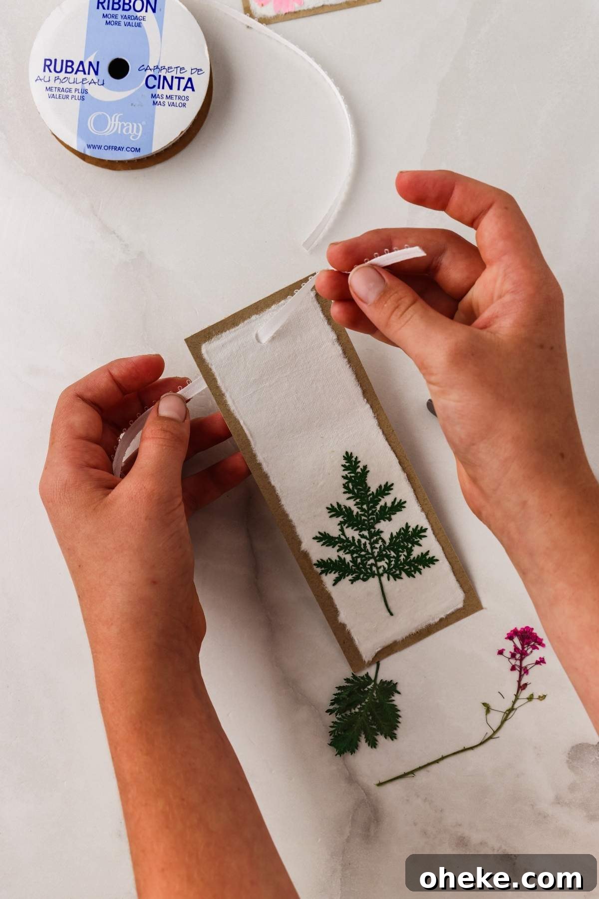 DIY Pressed Flower Bookmarks: Step-by-Step Craft Tutorial 18 Hands assembling a card with a pressed green leaf, reminiscent of creating pressed flower bookmarks. A ribbon spool and pink flower add charm nearby on the white surface.