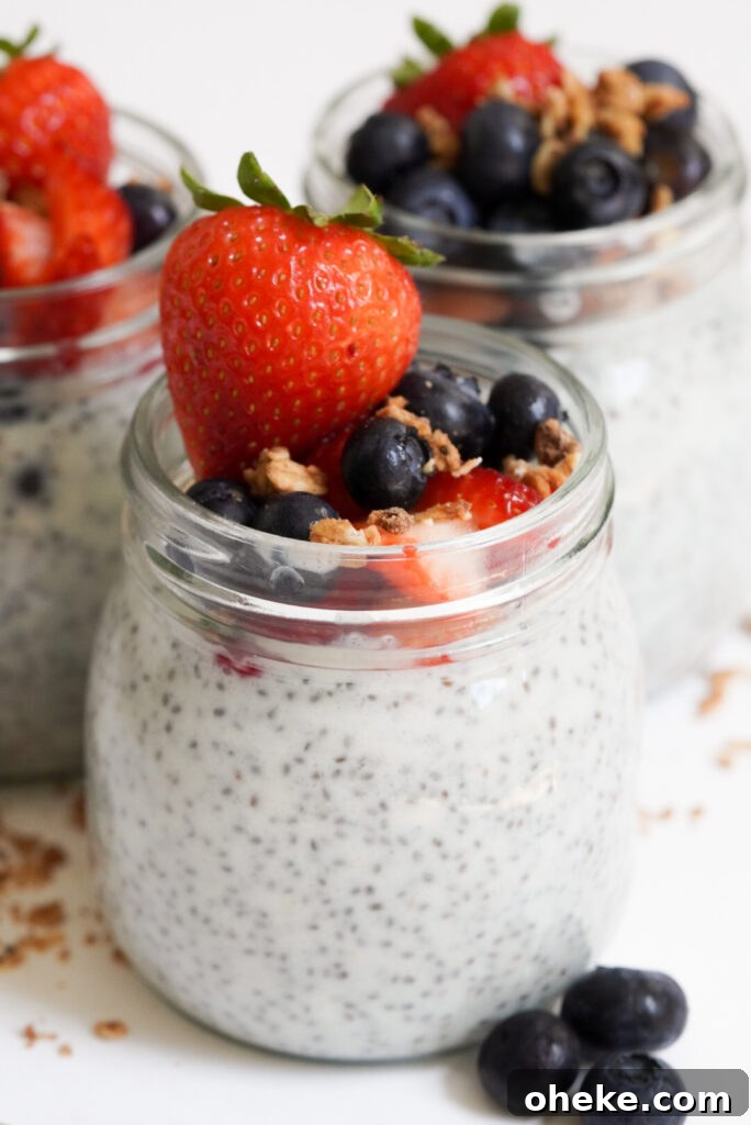 High-Protein Chia Pudding Recipe for Muscle Fuel 9 45 degree angle of 3 jars of high protein chia pudding on a white table with granola and blueberries scattered around.