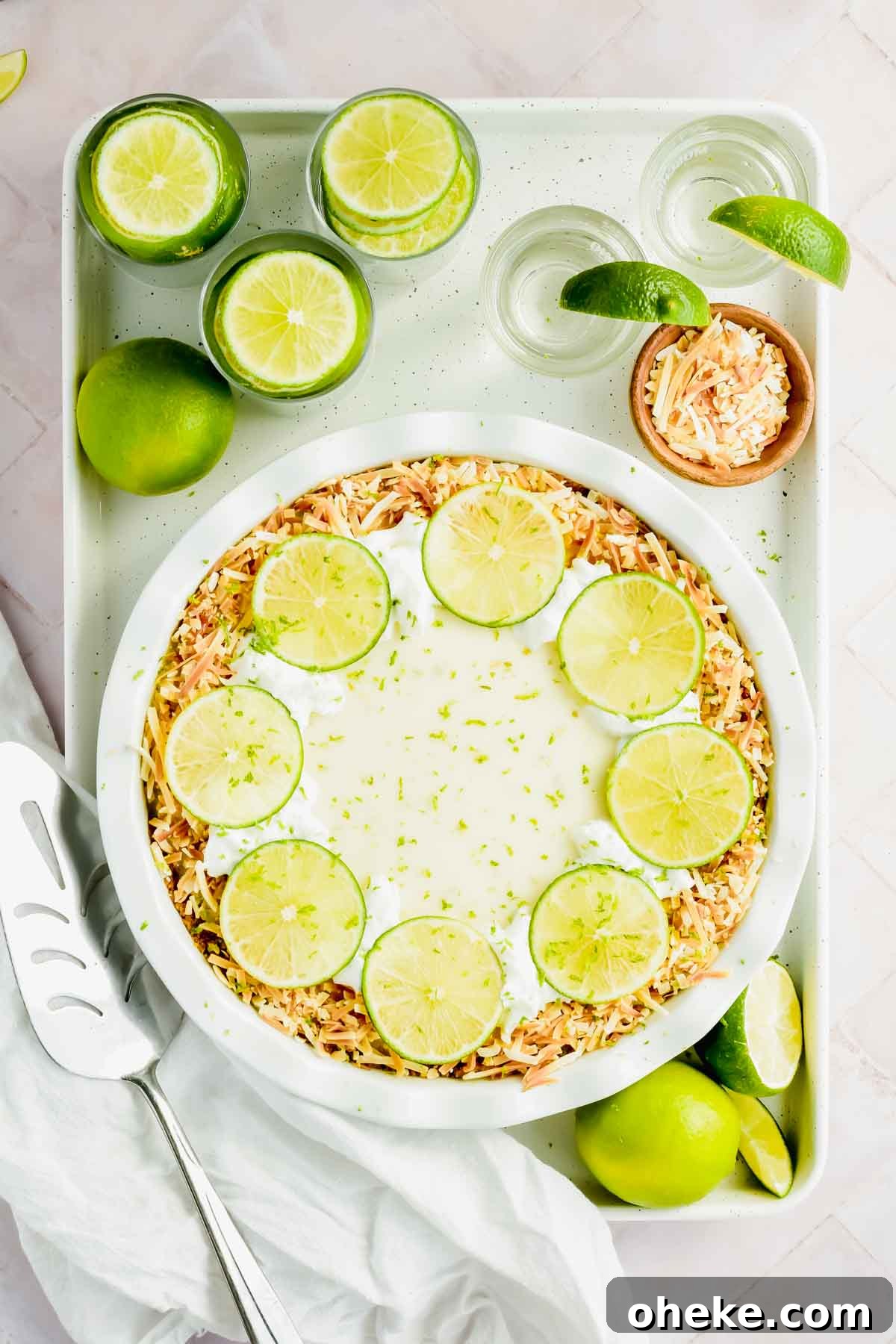 Tequila Lime Key Lime Pie Recipe with a Boozy Graham Cracker Crust 7 key lime pie garnished with toasted coconut and lime slices on white baking sheet surrounded by limes and tequila shots with white background.
