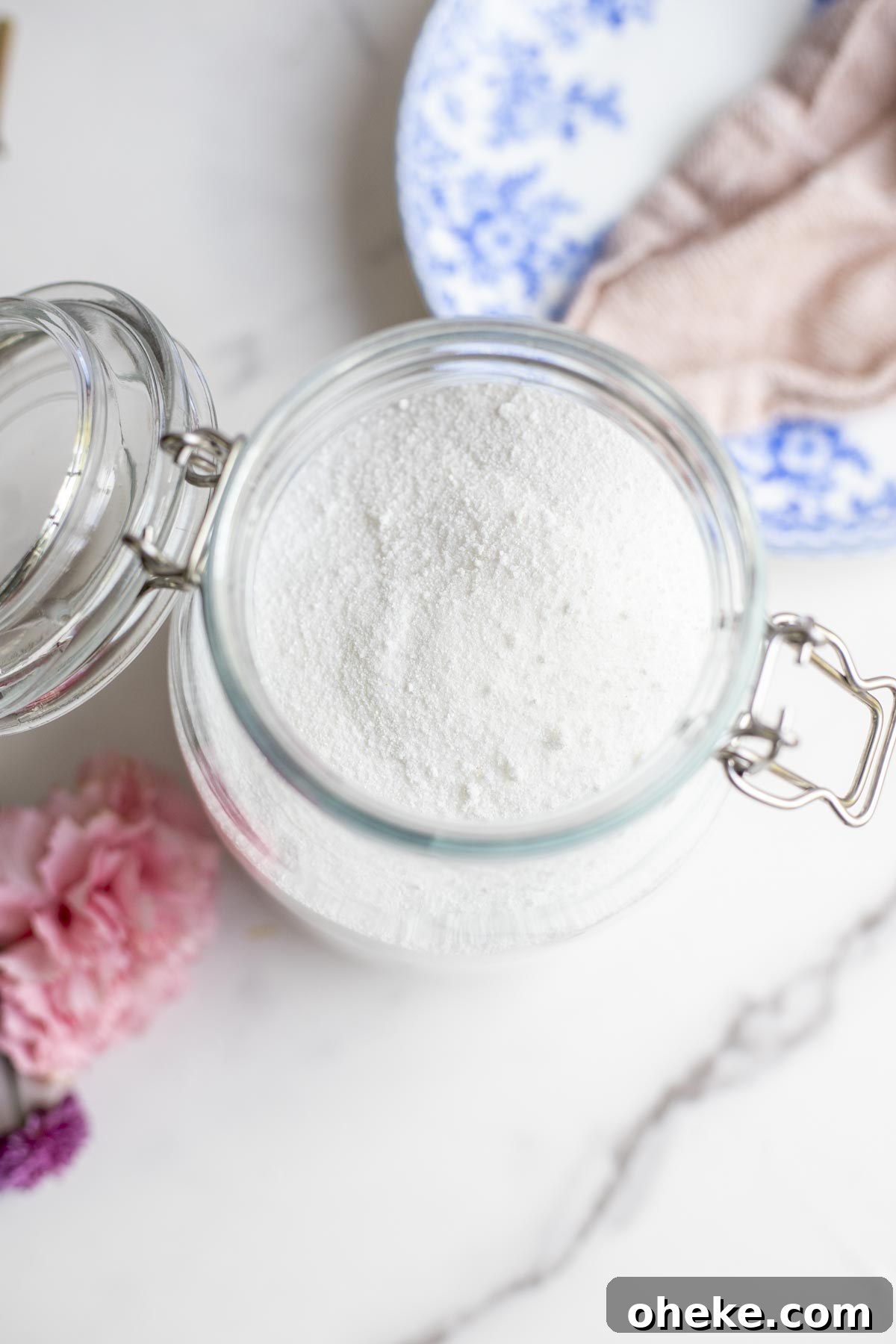 Overhead photo of dishwasher detergent in a glass swing top jar with a stack of dishes with a pink towel to the right.