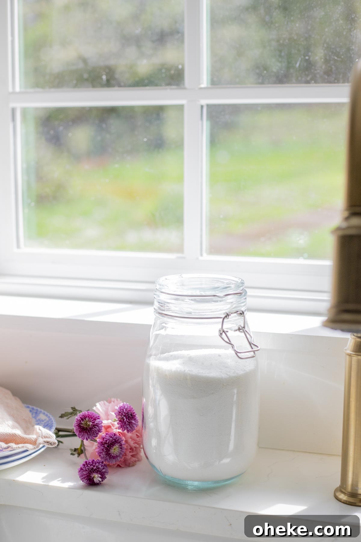Homemade dishwasher detergent in a glass container on a windowsill with flowers to the left.