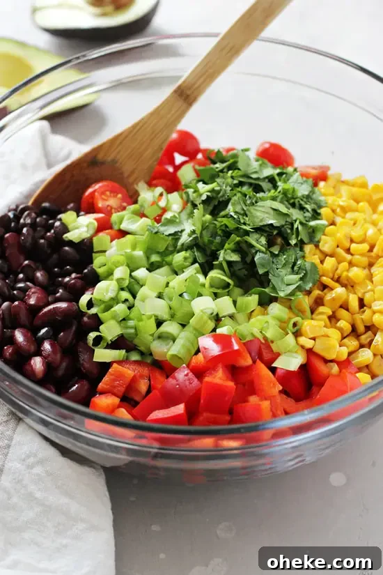 A glass bowl filled with all the components for Southwestern Bean Salad with a wooden spoon in the dish.