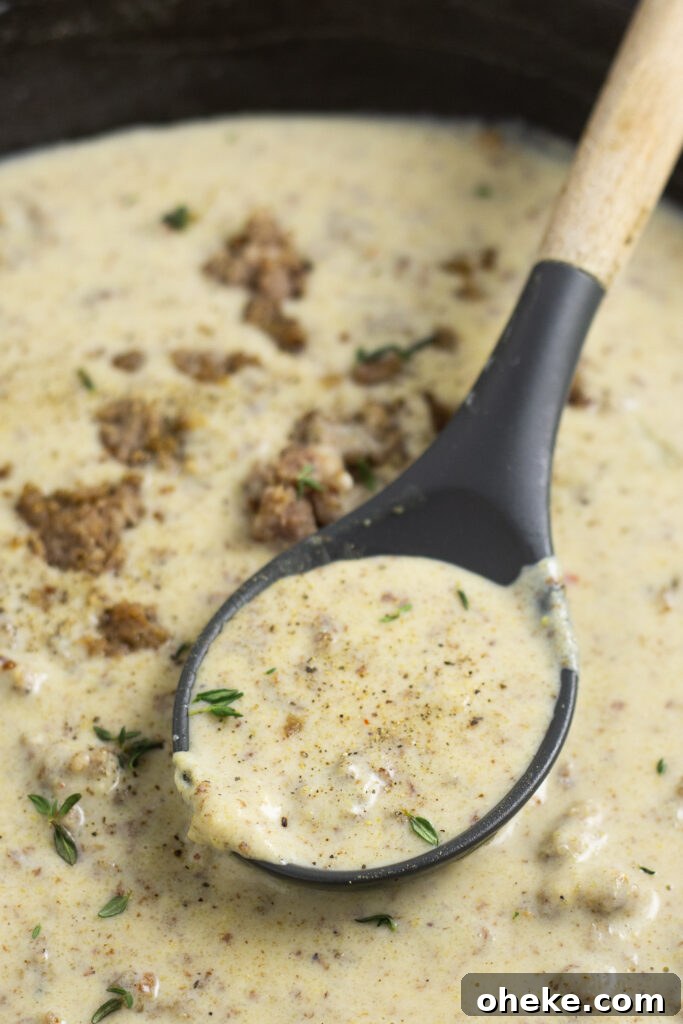 Cast iron skillet full of sawmill gravy topped with crumbled pork sausage and fresh thyme with a wooden spoon in it