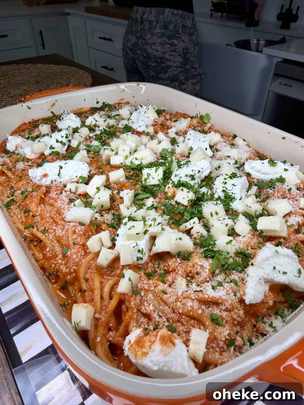 Creamy Italian Baked Spaghetti Recipe — a Tastier Twist on Baked Ziti 11 A large orange baking dish filled with spaghetti in red sauce, topped with chunks of white cheese, ricotta, and chopped herbs, sits on a kitchen counter. A person stands in the background near a stove and sink.