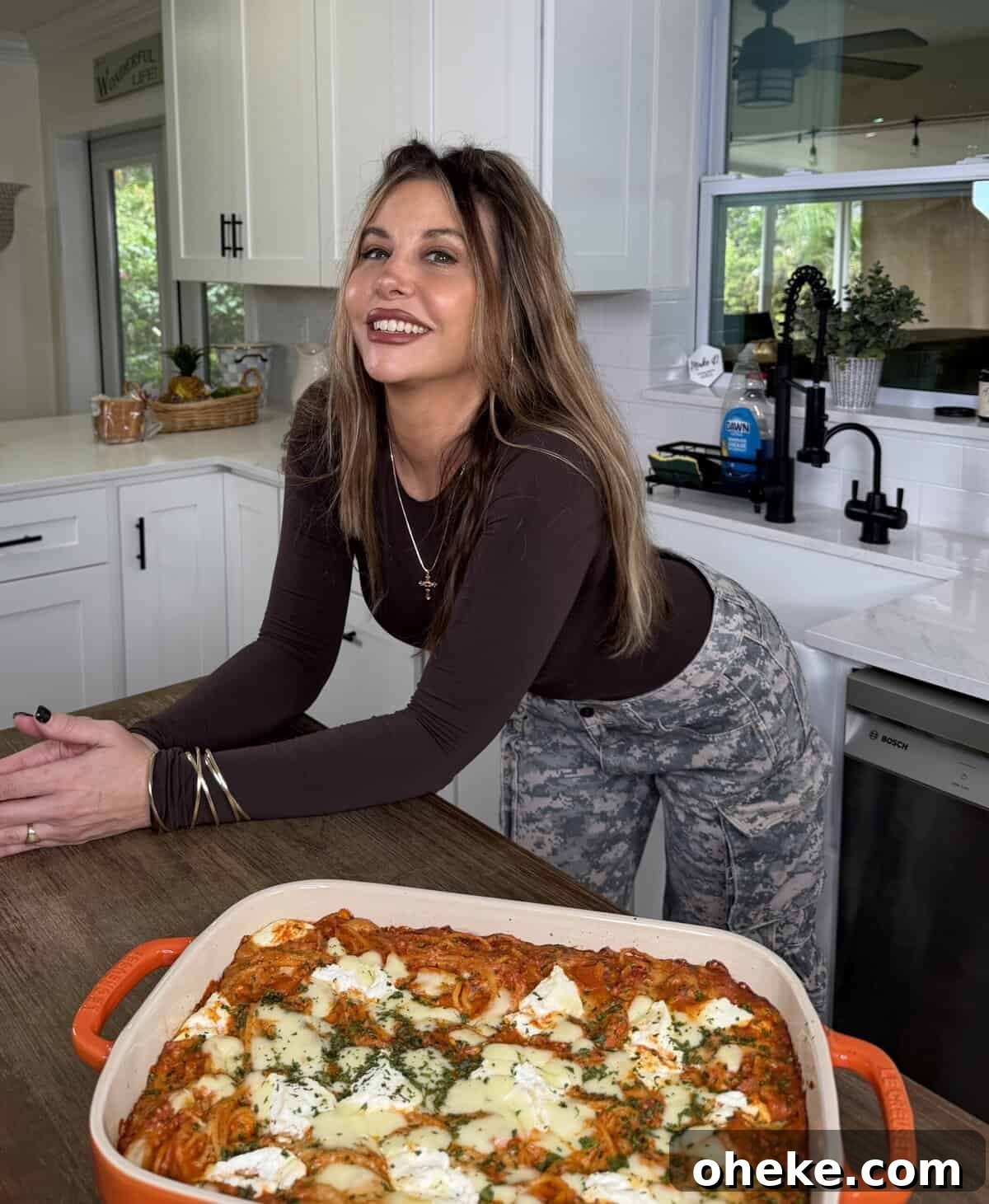 Creamy Italian Baked Spaghetti Recipe — a Tastier Twist on Baked Ziti 12 A woman with long brown hair smiles while leaning on a kitchen counter, wearing a dark long-sleeve top and camouflage pants. In front of her is a baked dish topped with melted cheese and herbs.