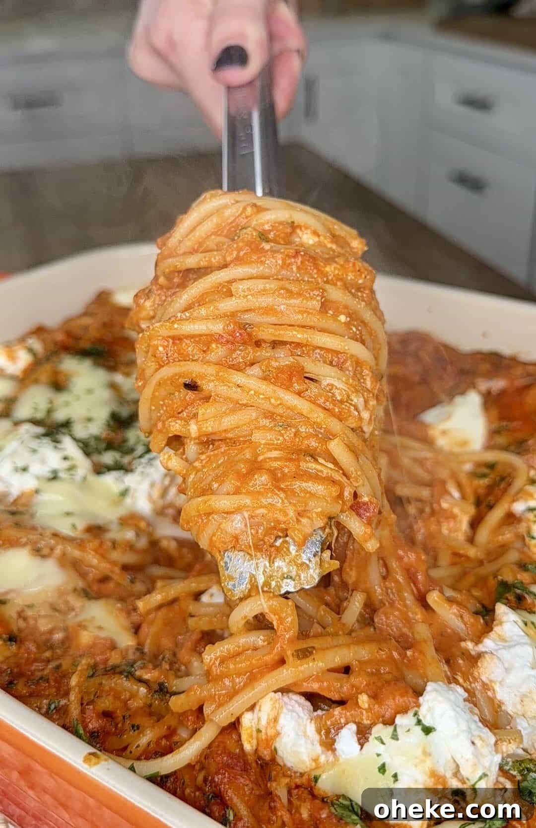 Creamy Italian Baked Spaghetti Recipe — a Tastier Twist on Baked Ziti 13 A hand holding tongs lifts a serving of cheesy baked spaghetti with tomato sauce from a casserole dish, with steam rising and melted cheese visible throughout.