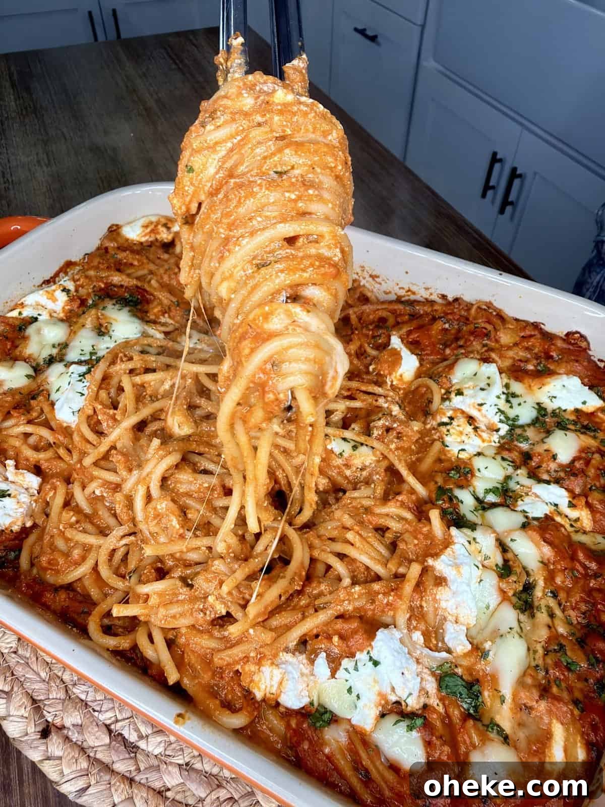 Creamy Italian Baked Spaghetti Recipe — a Tastier Twist on Baked Ziti 14 A close-up of spaghetti baked in tomato sauce with melted cheese and herbs in a casserole dish. A serving of spaghetti is being lifted with tongs, showing stretchy cheese. White cabinets are visible in the background.