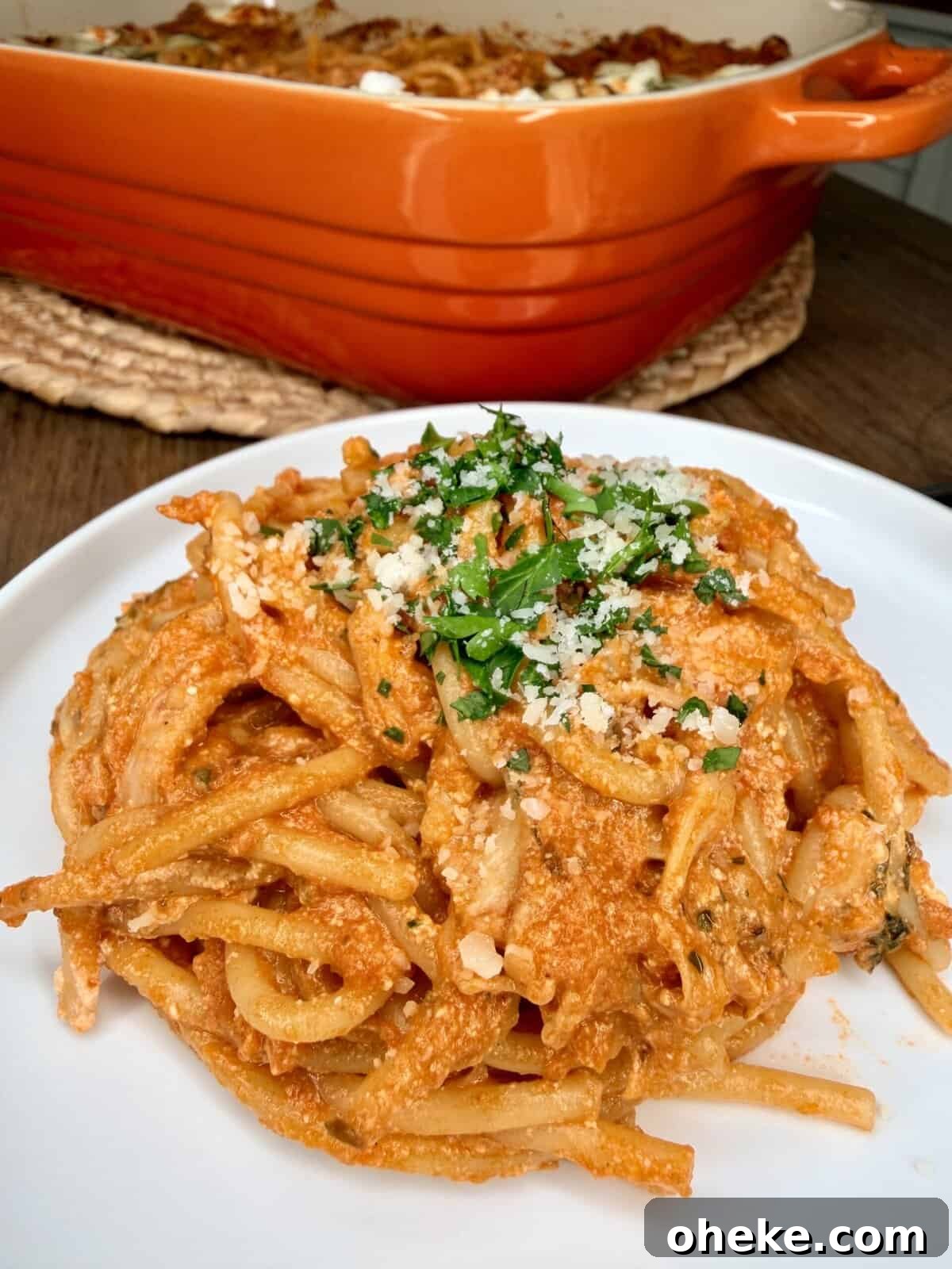 Creamy Italian Baked Spaghetti Recipe — a Tastier Twist on Baked Ziti 15 A plate of baked spaghetti with red sauce, cheese, and herbs sits in the foreground, garnished with grated cheese and parsley. In the background, a large orange baking dish filled with more pasta is visible.