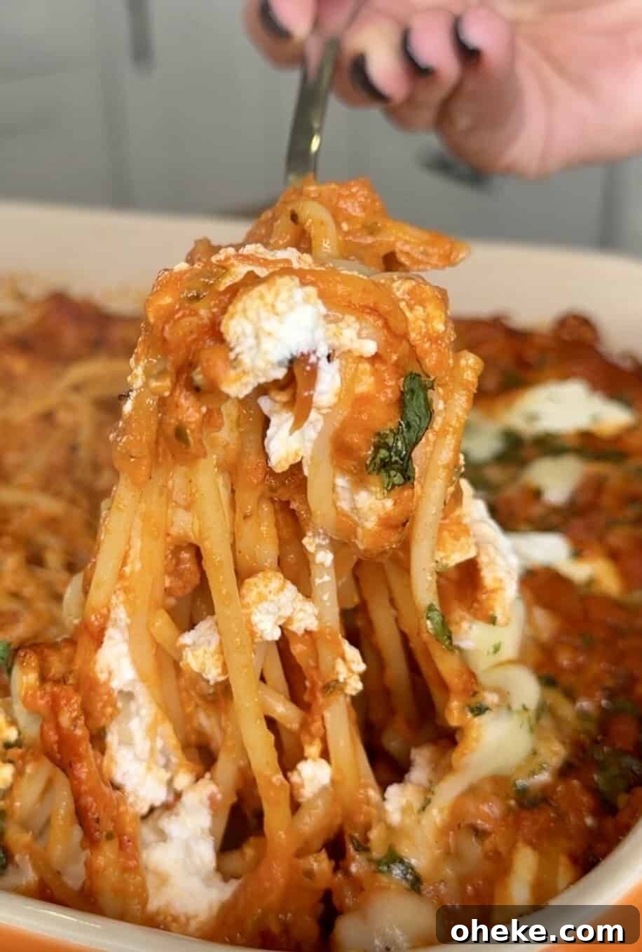 Creamy Italian Baked Spaghetti Recipe — a Tastier Twist on Baked Ziti 16 A hand holding a fork lifts a serving of baked spaghetti with tomato sauce, melted cheese, and herbs from a casserole dish.