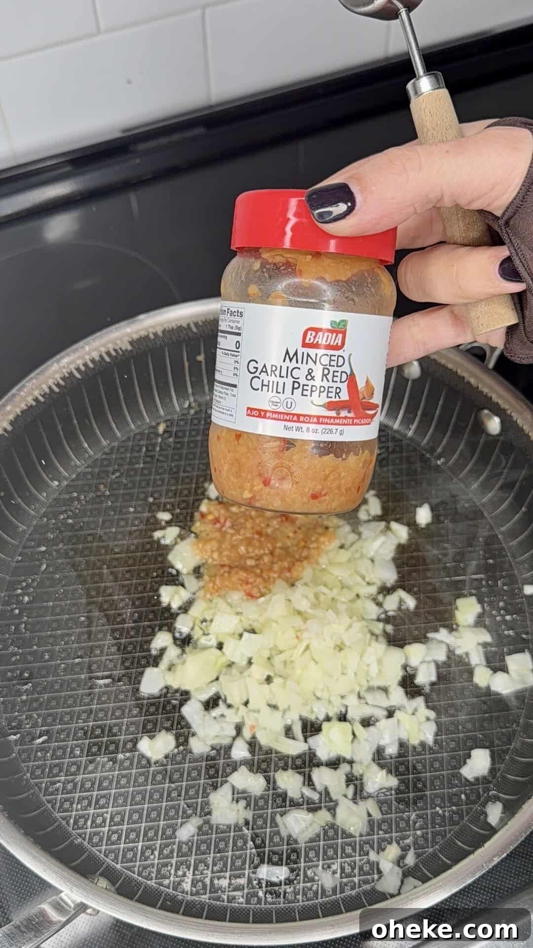 Creamy Italian Baked Spaghetti Recipe — a Tastier Twist on Baked Ziti 3 A hand holds a jar of Badia Minced Garlic & Red Chili Pepper over a pan with chopped onions sautéing on a stovetop.