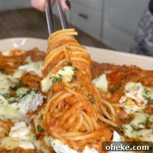 Creamy Italian Baked Spaghetti Recipe — a Tastier Twist on Baked Ziti 18 A hand uses tongs to lift a serving of cheesy baked spaghetti with tomato sauce and herbs from a casserole dish in a kitchen setting.