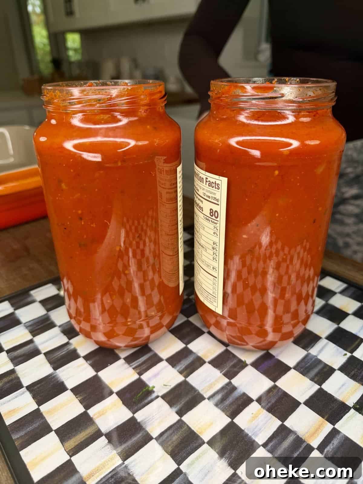 Creamy Italian Baked Spaghetti Recipe — a Tastier Twist on Baked Ziti 4 Two glass jars filled with vibrant red tomato sauce are placed side by side on a checkered tray in a kitchen setting. The jars have labels with nutritional information visible.