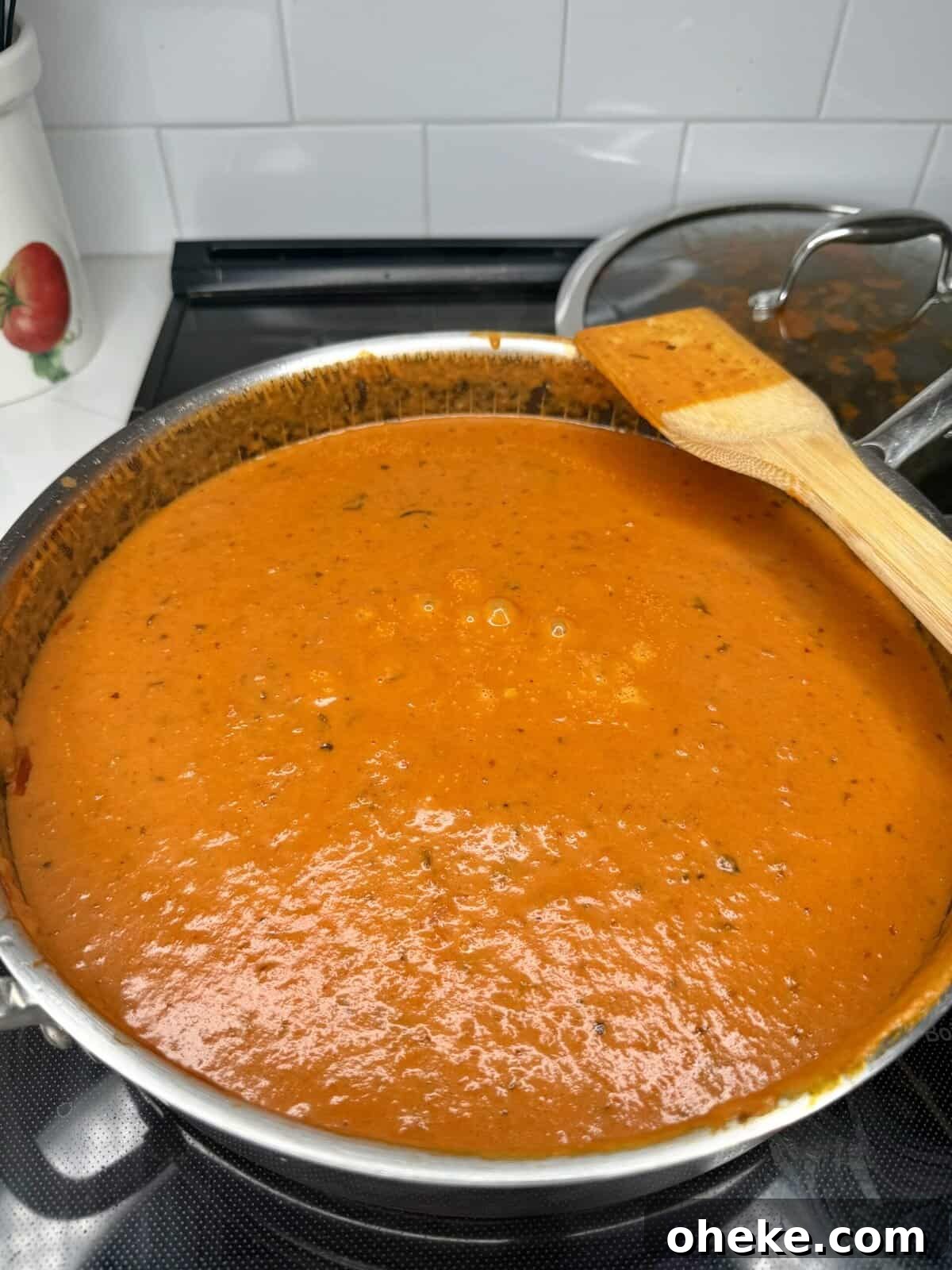 Creamy Italian Baked Spaghetti Recipe — a Tastier Twist on Baked Ziti 7 A large metal pot filled with creamy tomato soup sits on a stovetop. A wooden spatula rests across the side of the pot. A glass-lidded pot and kitchen decor are visible in the background.