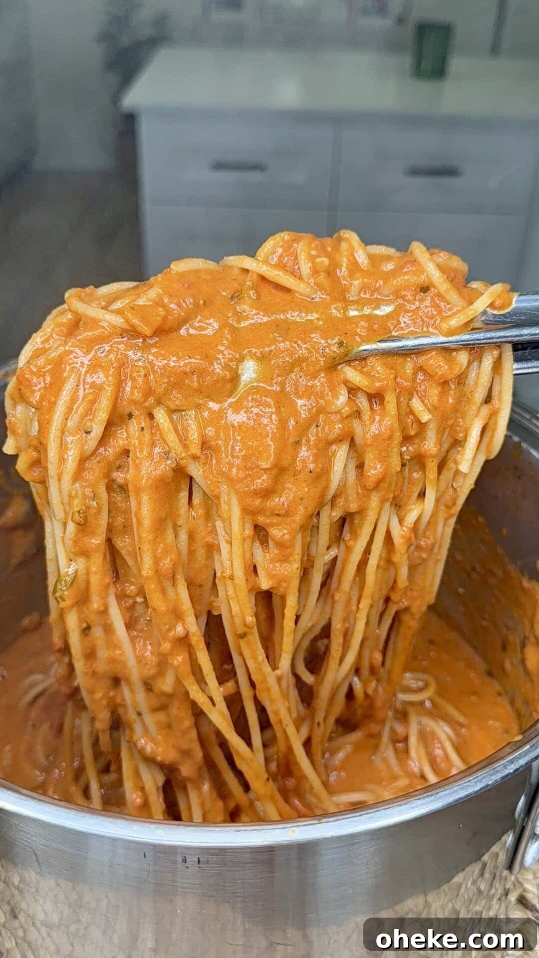 Creamy Italian Baked Spaghetti Recipe — a Tastier Twist on Baked Ziti 9 A close-up of spaghetti being lifted from a pot with tongs, coated in a thick, creamy tomato sauce. A white kitchen is slightly visible in the blurry background.