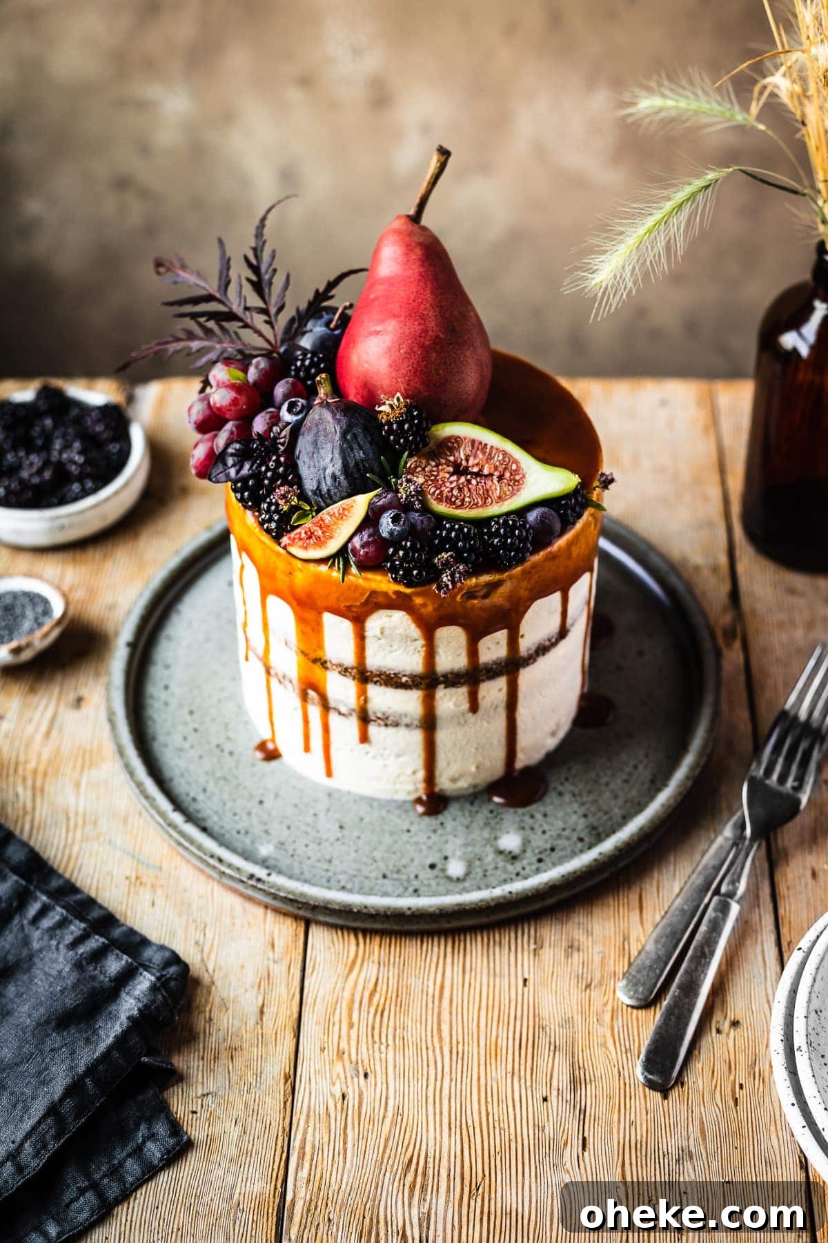 Blackberry Poppy Seed Bundt Cake with Lemon Glaze 2 A 45 degree angle view of a layer cake with a caramel drip and a crown of late summer fruit on top. The cake sits on a blue grey ceramic platter on a rustic wooden table with forks, a dark blue linen napkin, and small bowls of poppy seeds and blackberries nearby. There is a warm tan stone background behind the table.