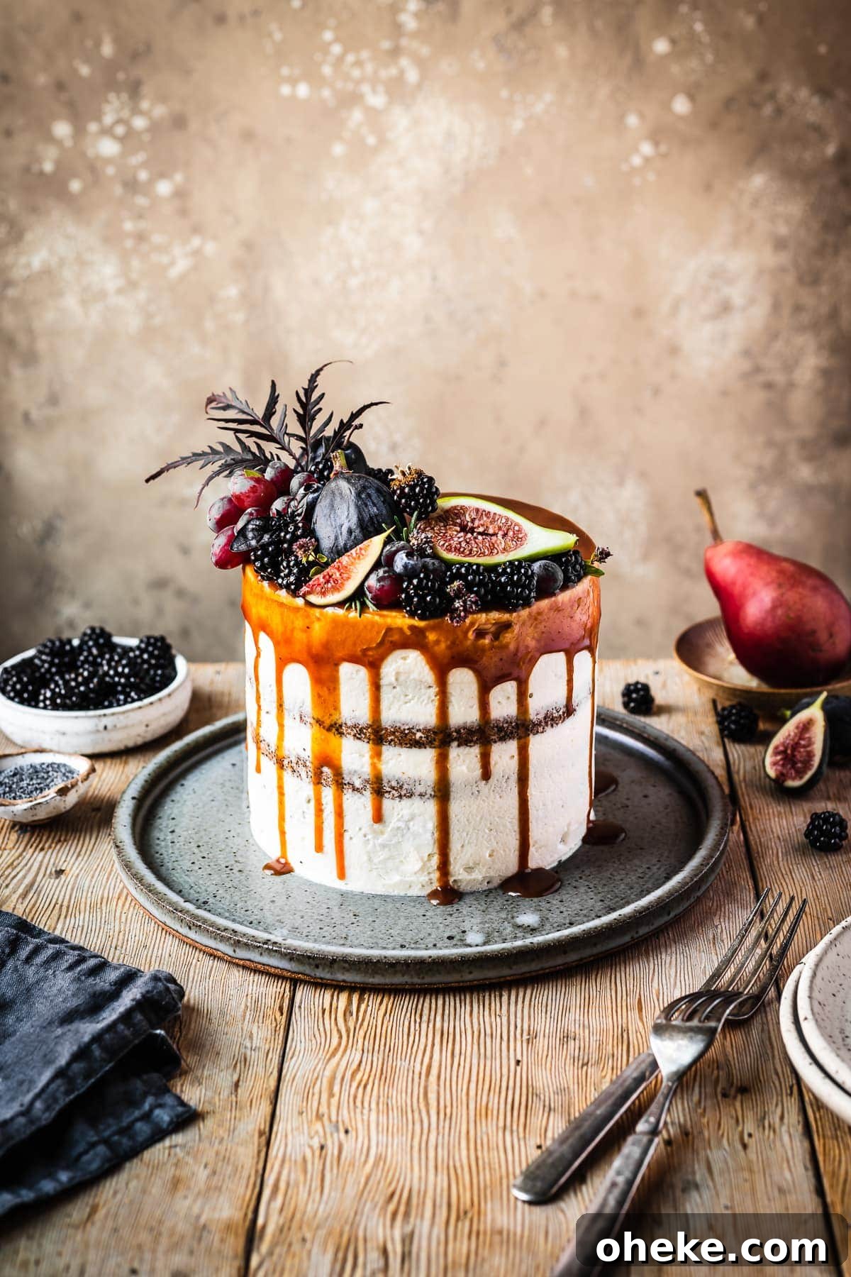 Blackberry Poppy Seed Bundt Cake with Lemon Glaze 11 A white frosted blackberry poppy seed layer cake with a caramel drip and a crown of late summer fruit on top. The cake sits on a blue grey ceramic platter on a rustic wooden table with forks and small bowls of poppy seeds and blackberries nearby. There is a warm tan stone background behind the table.