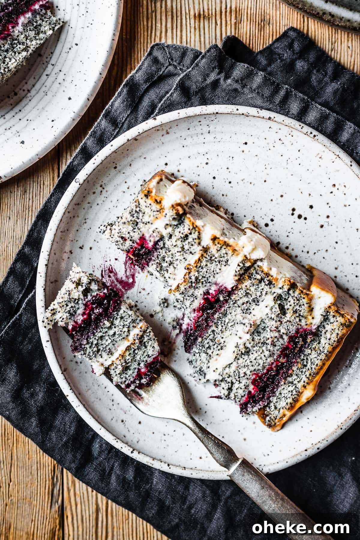 Blackberry Poppy Seed Bundt Cake with Lemon Glaze 3 A slice of poppyseed cake with mascarpone frosting and caramel drip, with layers of vibrant purple blackberry basil jam. The slice sits on a white speckled ceramic plate with a navy blue linen napkin folded underneath, on a rustic wooden table.