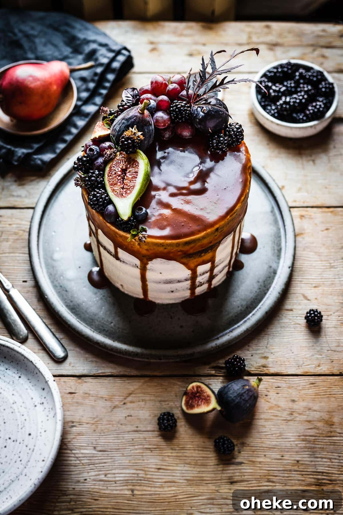 Blackberry Poppy Seed Bundt Cake with Lemon Glaze 5 A white frosted layer cake with a caramel drip and a crown of late summer fruit on top. The cake sits on a blue grey ceramic platter on a rustic wooden table with forks and small bowls of poppy seeds and blackberries nearby. The light is coming from behind the cake and makes the caramel on top of the cake shine.
